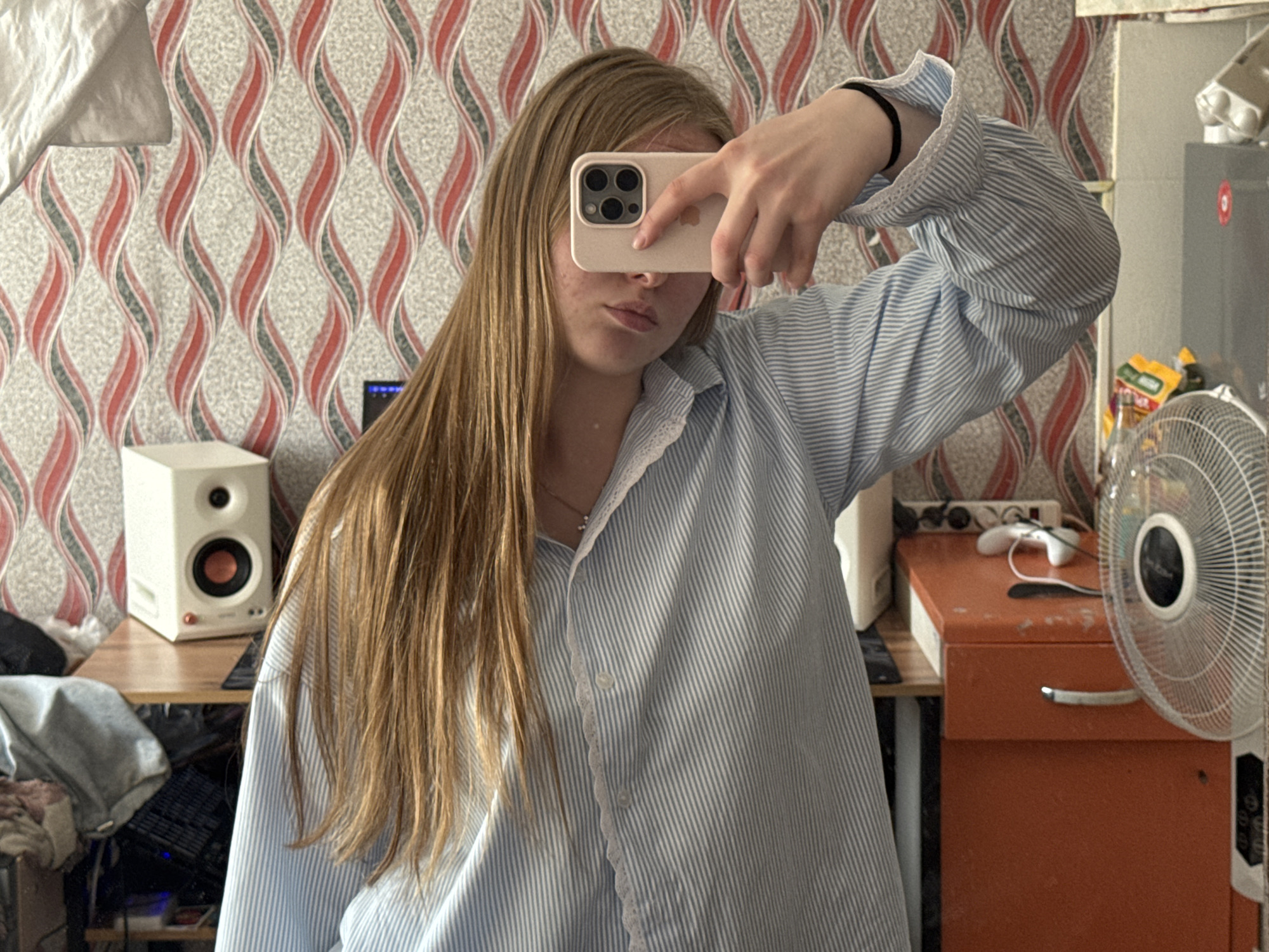 A person is taking a selfie indoors. The person has long, light brown hair and is wearing a light blue and white striped shirt. The phone is held in front of their face, obscuring part of their features.
In the background, there is a wall with a patterned wallpaper in red, gray, and white. To the left, there is a white speaker and a desk with various objects on it. To the right, there is a gray refrigerator and an orange storage unit. A white fan is partially visible in the right foreground.
Based on the appearance of the room, the objects in it, and the Russian text on the packaging, the location is most likely Perm, Russia.