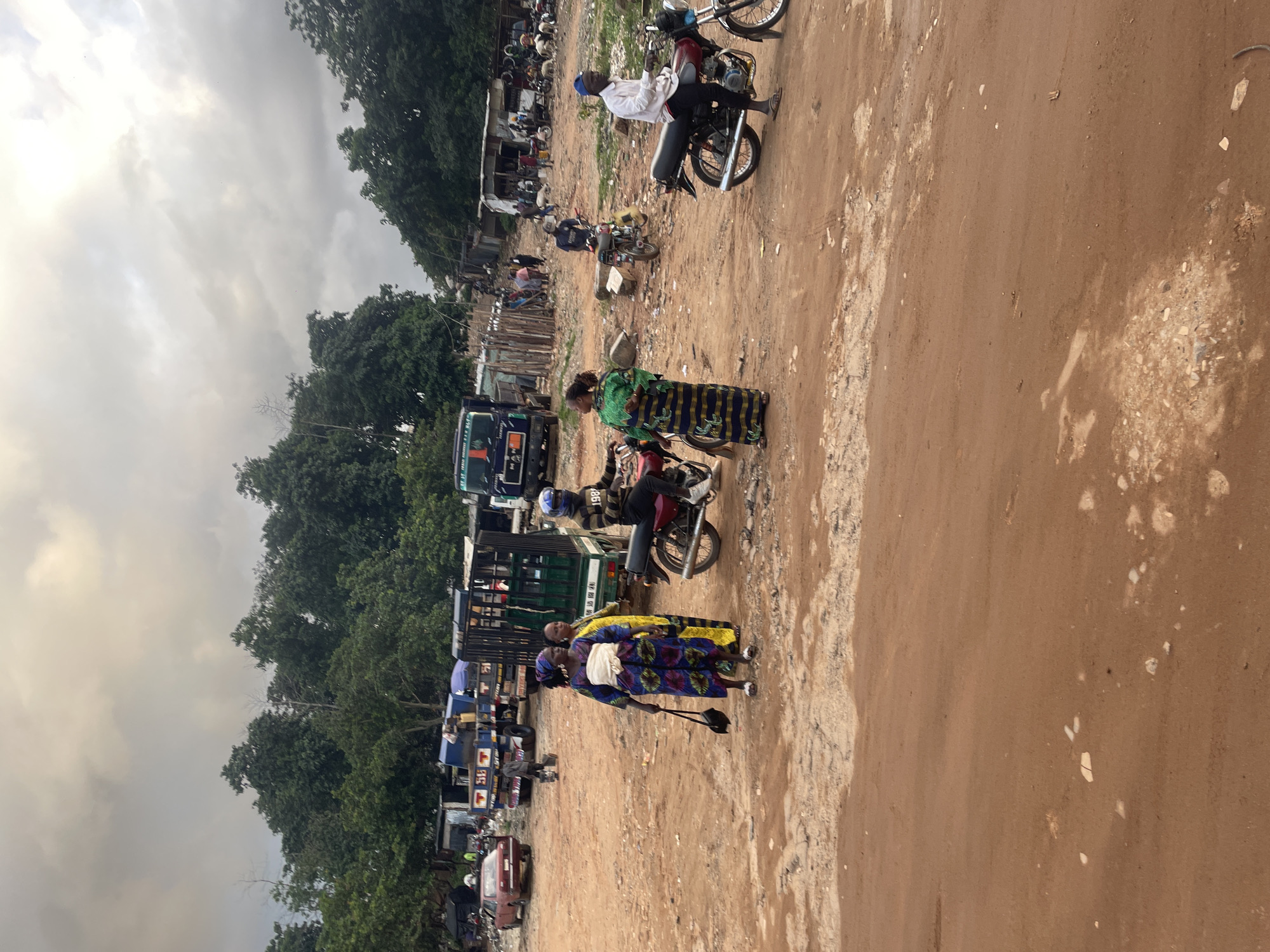 This is a forensic, matter-of-fact description of the image, set in the city of Kurunduma, Nigeria.
The image depicts a dusty, outdoor marketplace or transportation hub with a cloudy sky overhead. Several vehicles, including trucks, cars, and motorcycles, are present. In the foreground, two women in traditional African attire are walking. One woman, in a blue and purple patterned dress with a white sash, carries a dark handbag. The other woman, in a yellow and green patterned dress, stands beside her. To the right, a man wearing a white long-sleeved shirt, dark pants, and a blue cap rides a red motorcycle. Another man in a black and yellow striped jacket and blue helmet rides a similar motorcycle in the middle ground. A green truck with a caged rear is parked behind the women. Further in the background, more trucks, market stalls, and scattered debris are visible. The ground is predominantly reddish-brown dirt with scattered stones and patches of lighter colored earth. The overall scene suggests a busy, informal commercial area.