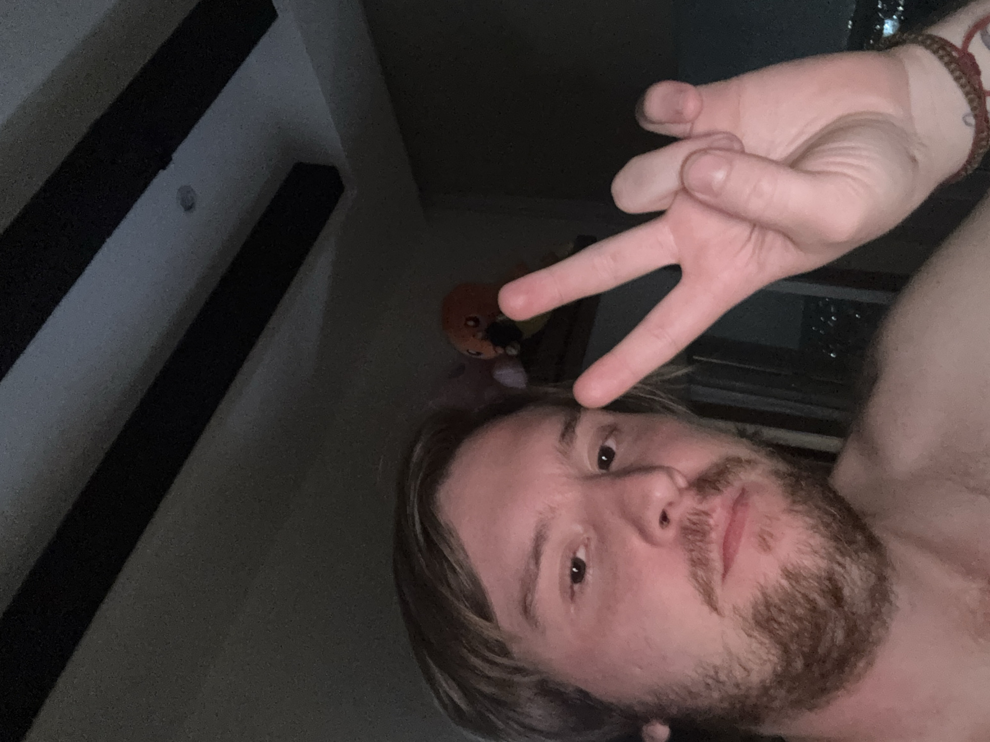 A selfie taken indoors in low light depicts a fair-skinned person with light brown hair and a beard. They are holding their hand up, making a "peace" or "victory" sign. A bracelet is visible on their wrist. A small orange plush object sits in the background. Details regarding the exact location in Huixquilucan de Degollado, Mexico, are not discernible from the image.