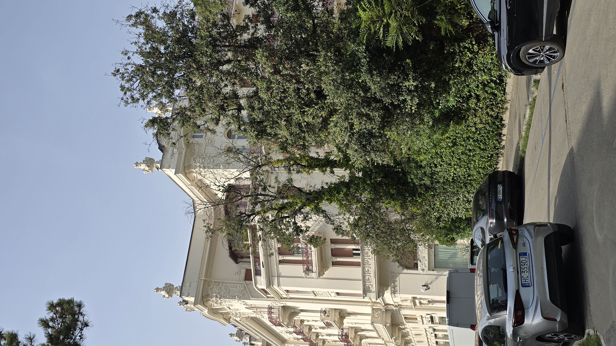 The image depicts the facade of the Grand Hotel Rimini, a historic and iconic hotel located in Rimini, Italy. The hotel is a magnificent building with ornate architectural details, featuring balconies, elaborate carvings, and classical embellishments. Lush green trees and foliage surround the hotel, partially obscuring its lower levels and creating a picturesque contrast with the cream-colored architecture. In the foreground, several cars are parked along the street, including a silver MG and a black Porsche. The scene is illuminated by natural daylight, suggesting it was taken during the day. The overall impression is one of elegance and grandeur, typical of a high-end establishment in a coastal Italian city.