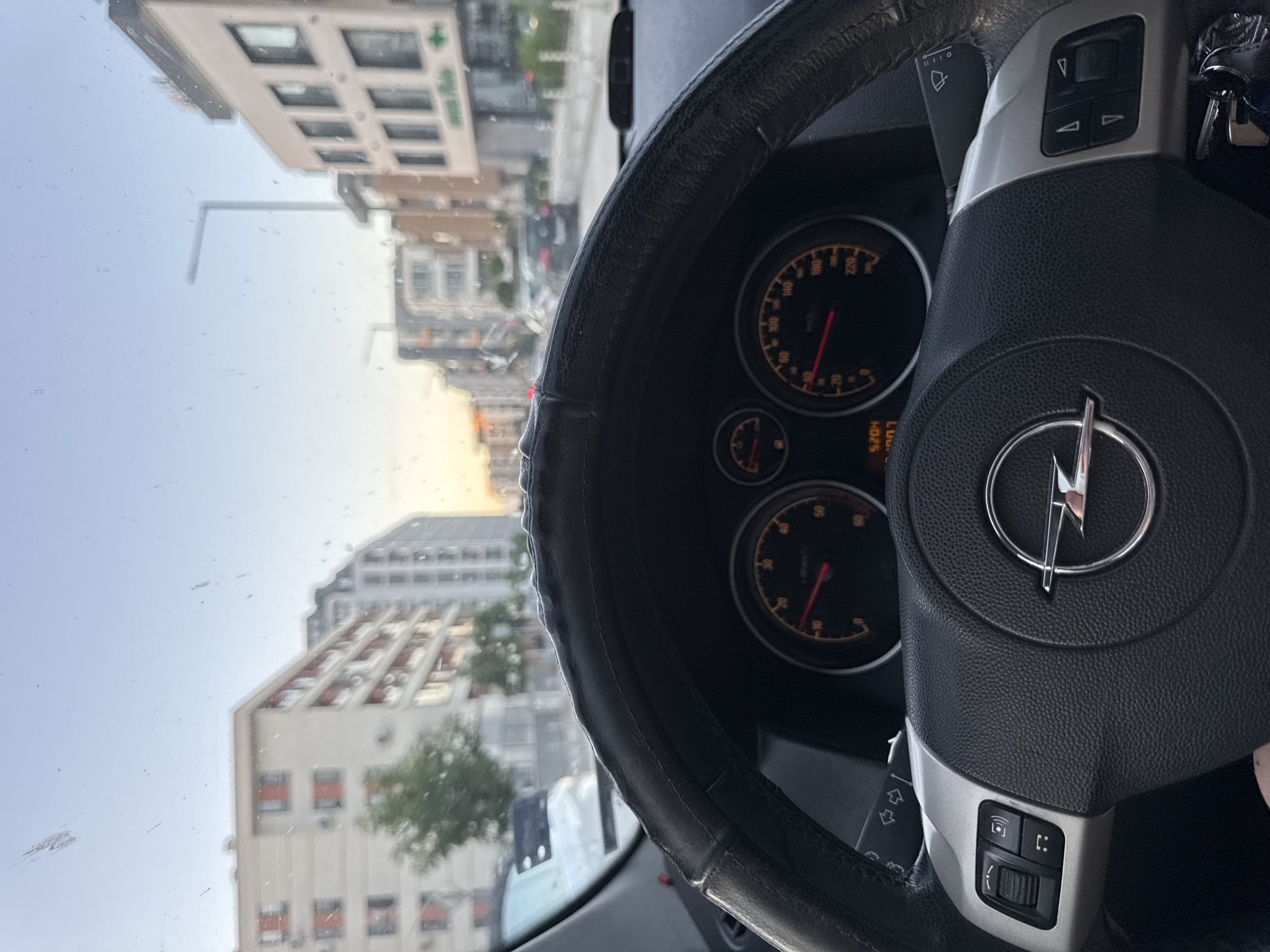 The image shows the interior of a car, specifically focusing on the steering wheel with the Opel logo. The speedometer is visible, displaying 5204km. Through the windshield, an urban landscape is visible, including buildings and a street. The location is Sofia, Bulgaria, as evidenced by the distinct architecture and urban layout characteristic of the city. The image is captured during daylight, with clear sky.