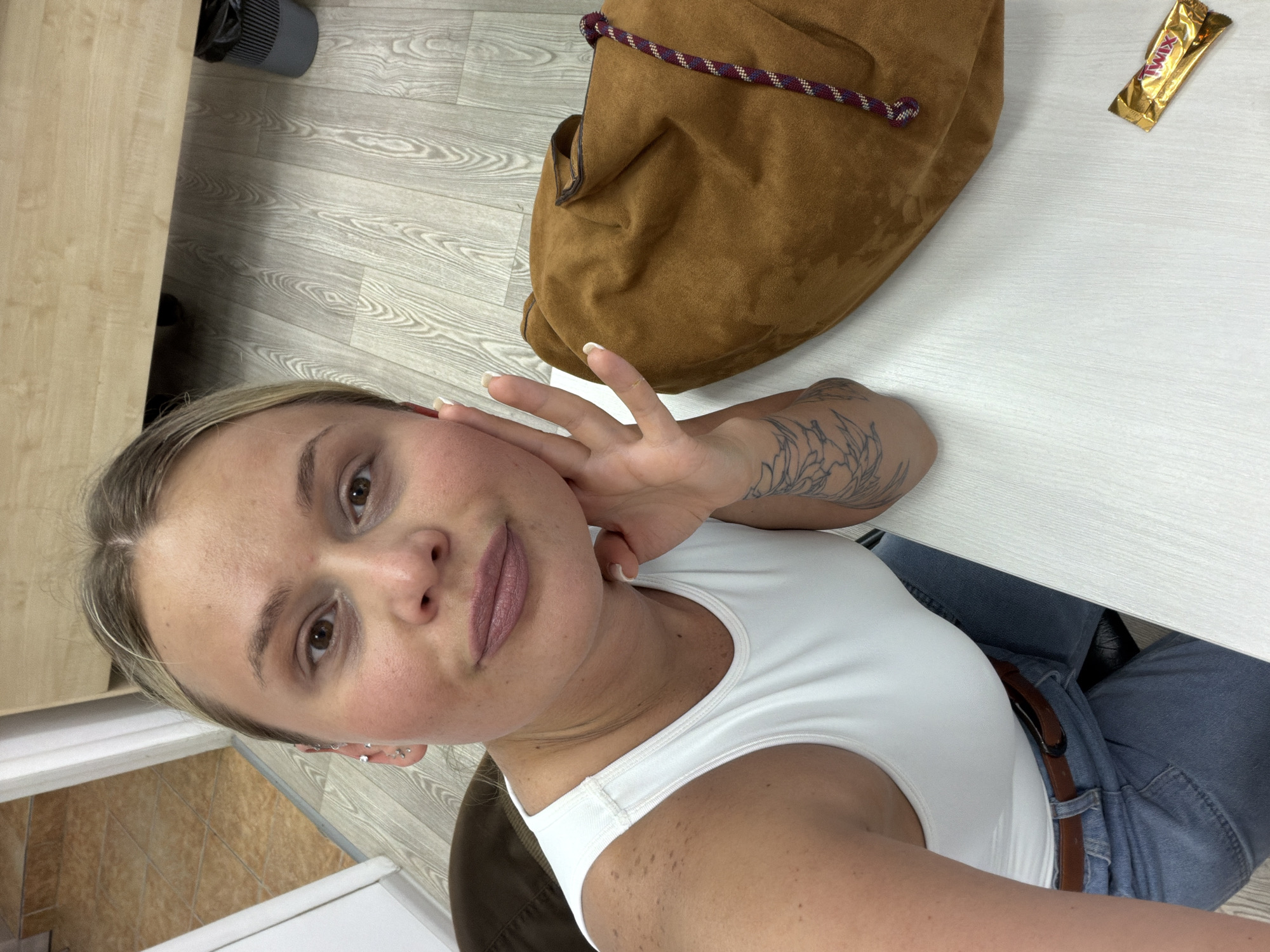 A woman is in a room with light wood flooring and a white desk, taking a selfie. She has fair skin, light brown hair, and a tattoo on her forearm. She is wearing a white tank top and blue jeans with a brown belt. A brown suede bag with a drawstring is on the desk, as is a Twix candy bar. There is a gray air purifier in the background. The location is potentially Sarapul, Russia.