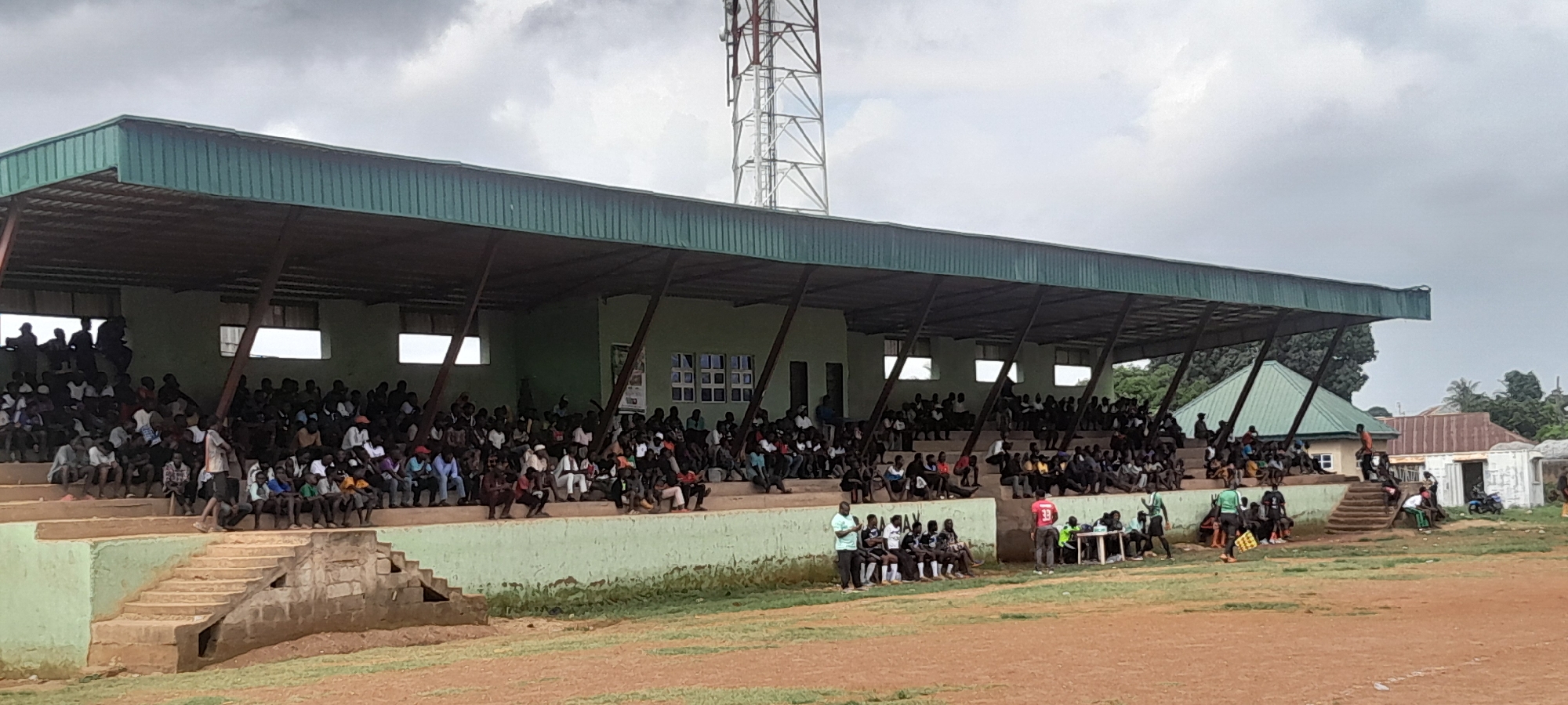 The image depicts a partially filled outdoor stadium seating area, likely within Abuja, Nigeria, given its architecture and setting. The stadium features a green, corrugated metal roof supported by dark metal beams, providing shade over tiered concrete seating. A large, silver lattice telecommunications tower is prominently visible in the upper right quadrant of the image, extending above the stadium structure and trees. A crowd of spectators, predominantly men and boys, are seated on the concrete tiers, some appearing to be engaged in conversation while others watch an event off-camera. In the foreground, a dirt field and grassy patches indicate the presence of a sports ground. Several individuals are gathered near the bottom of the seating area, some appearing to be players or staff, with one person wearing a green jersey and shorts, and another in a red shirt. A table with chairs is set up near this group. The overall atmosphere suggests a local sporting event or gathering under an overcast sky. The surrounding area shows glimpses of buildings with corrugated roofs and dense green foliage, consistent with a suburban or less developed urban environment.