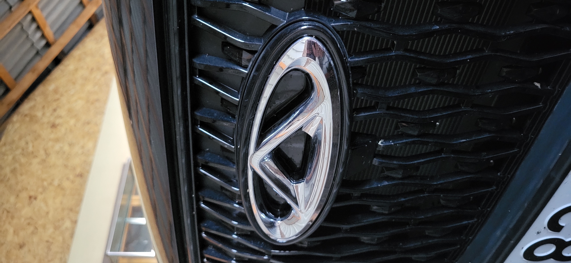 A close-up, slightly angled shot shows the chrome emblem of a Chery car, situated on its black grille. The emblem is a stylized oval with an internal design. The grille itself has a textured, wavy pattern. In the background, part of a car's black hood is visible, along with the wooden interior of a structure, possibly a garage or shed, with metal roofing and wooden beams overhead. A partial view of a license plate, featuring the numbers "82," is visible at the bottom of the frame. The image does not contain any identifiable features of the city of Taldykorgan, Kazakhstan.