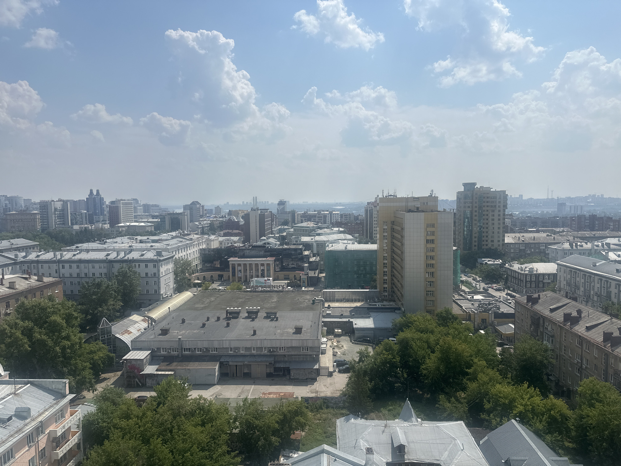 The image shows an aerial view of a cityscape in Novosibirsk, Russia, on a clear day with scattered clouds. The foreground features several buildings with flat, grey roofs and trees. A large, rectangular building with multiple air conditioning units is prominent in the center. To the right of this, a taller, multi-story apartment building with balconies is visible. In the background, a dense collection of various urban structures, including residential buildings, commercial properties, and industrial facilities, extends towards the horizon. A light haze softens the distant cityscape. The overall scene depicts a typical urban environment with a mix of architectural styles and green spaces.