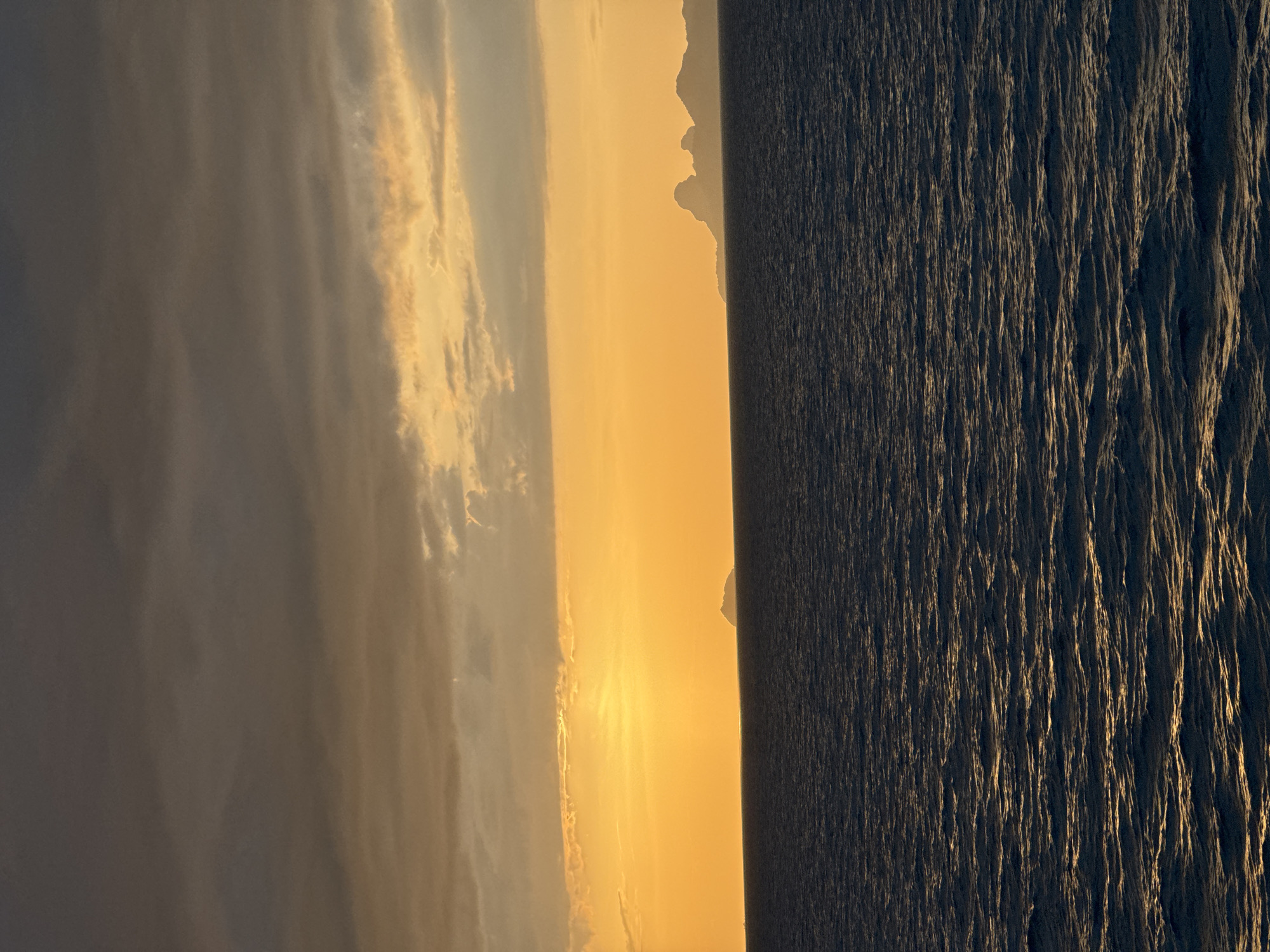 The image depicts a tranquil seascape at dusk. The sky is dominated by a gradient of colors, transitioning from a deep, moody gray at the top to a vibrant golden yellow near the horizon. Wispy clouds are scattered across the sky, catching the last rays of the setting sun.
In the distance, a chain of islands or a coastline is silhouetted against the bright sky. The closest island appears as a small, distinct shape, while further along the horizon, more complex mountainous landforms are visible, their peaks softened by the atmospheric haze.
The sea occupies the lower half of the image. It is a deep, dark blue, with gentle ripples creating a textured surface. The setting sun casts a warm, golden reflection on the water, creating streaks of light that shimmer across the waves.
There are no people or man-made objects visible in the scene. The environment is entirely natural, showcasing the beauty of a sunset over the ocean. The image conveys a sense of peace and serenity.