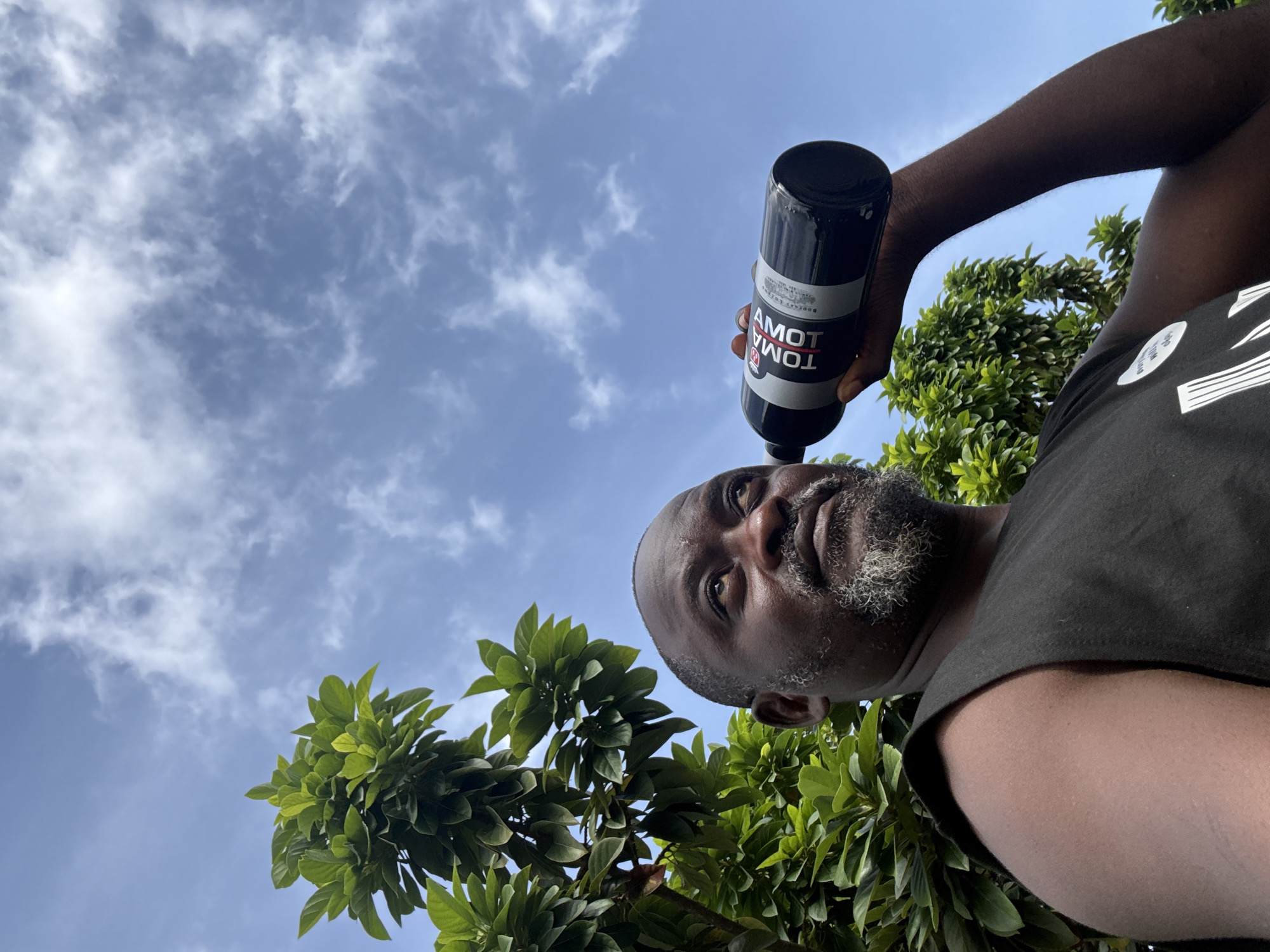 A low-angle shot shows a Black male holding a black bottle of TOMBOLA TOMATO brand product, likely in the city of Ado, Nigeria. The man, with a graying beard and wearing a black tank top, looks directly at the camera. Green trees surround the lower part of the frame. A blue sky with scattered white clouds is visible in the background.