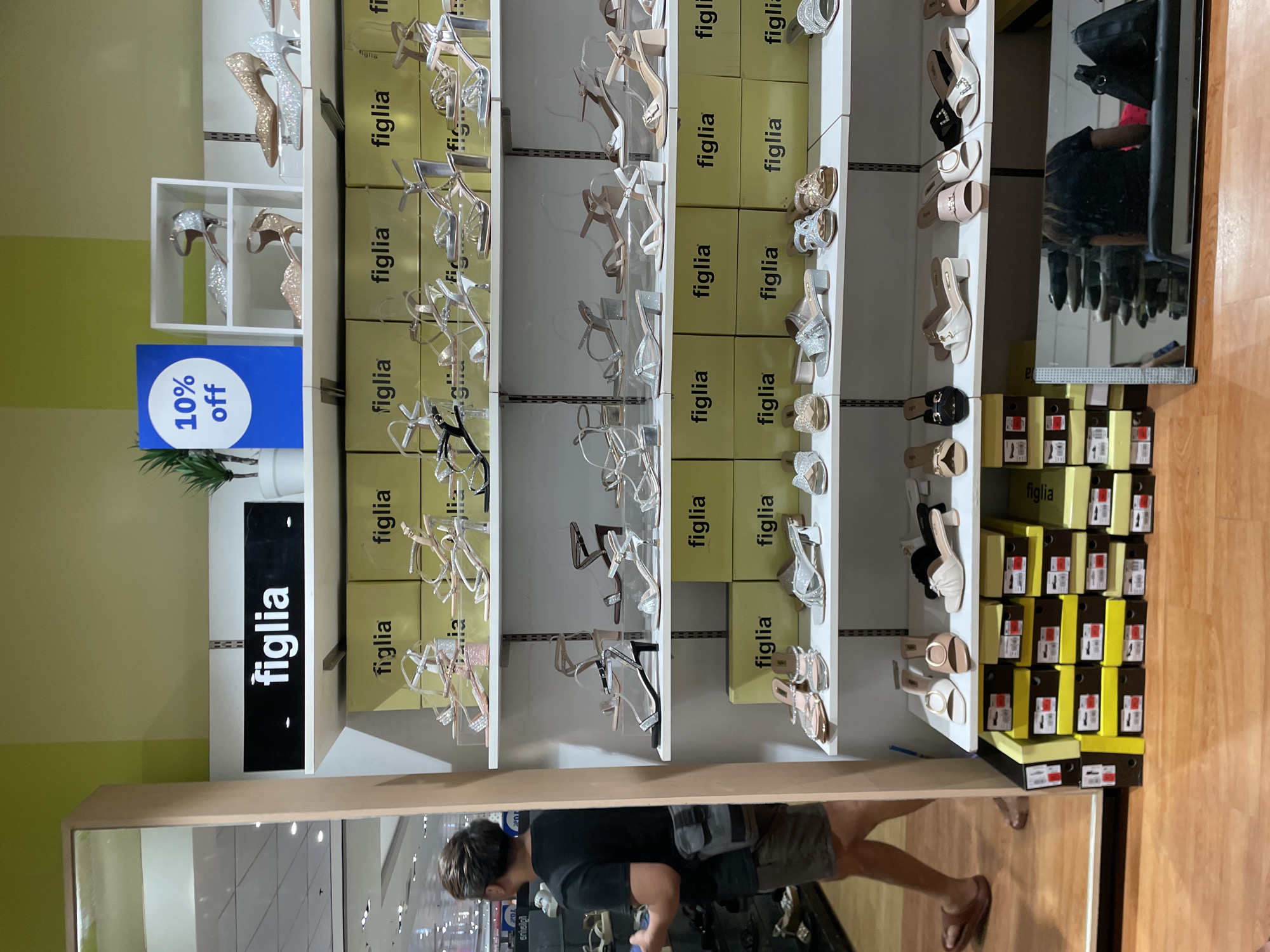 The image depicts an indoor scene within a Figlia shoe store located in Davao City, Philippines. The store features multiple white shelving units against a two-toned light green wall. Numerous pairs of women's shoes, including high heels, sandals, and mules, are displayed on these shelves. Many shoes exhibit glitter, metallic finishes, or clear straps, in colors such as silver, gold, pink, black, and white. Bright yellow shoeboxes, each prominently branded "figlia" in black text, are stacked behind and beneath the displayed shoes, forming the backdrop for several shelves. A black sign bearing the white "figlia" logo is mounted on the topmost shelf. Below it, a blue rectangular sign displays "10% off" in white text, positioned near a small potted artificial plant. Additional shoeboxes, some yellow and some darker, are stacked on the floor to the left. The floor is constructed of light-colored wood or laminate. On the left side of the frame, a large, vertical mirror reflects a male individual, seen from behind, walking towards the left. He has short, styled dark hair with highlights, wears a dark t-shirt, dark shorts, and brown slide sandals, and carries a gray cross-body bag. On the right, a horizontal mirror placed on the floor reflects a person, likely female, seated on a black bench, wearing a dark top, with a black handbag beside them.