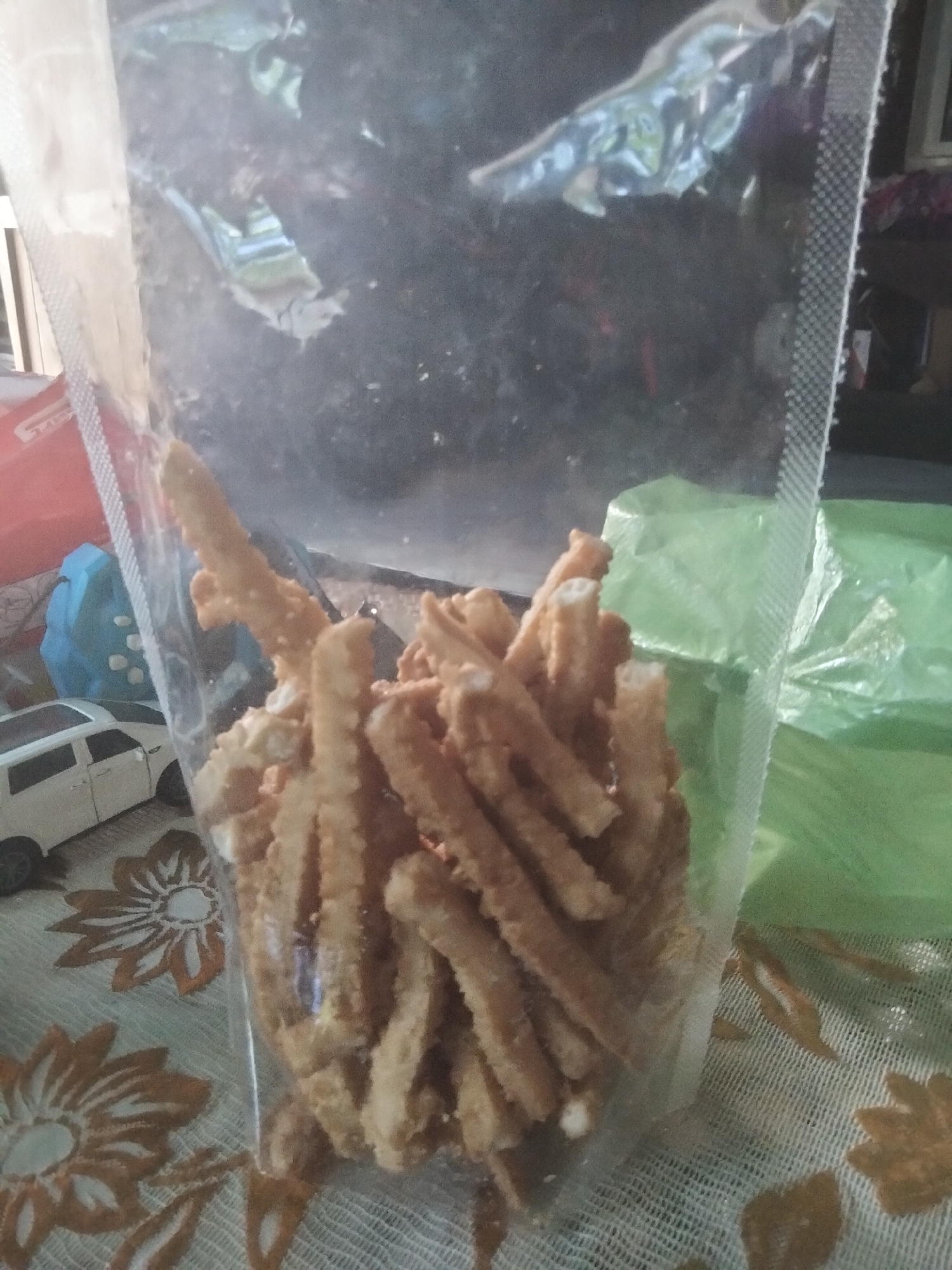 A clear plastic bag filled with brown, textured, stick-like snacks is sitting on a table with a floral patterned tablecloth. A toy car and a blue toy are visible beside the bag. A green plastic bag is behind the bag. The location is Kruengmanyang, Indonesia.