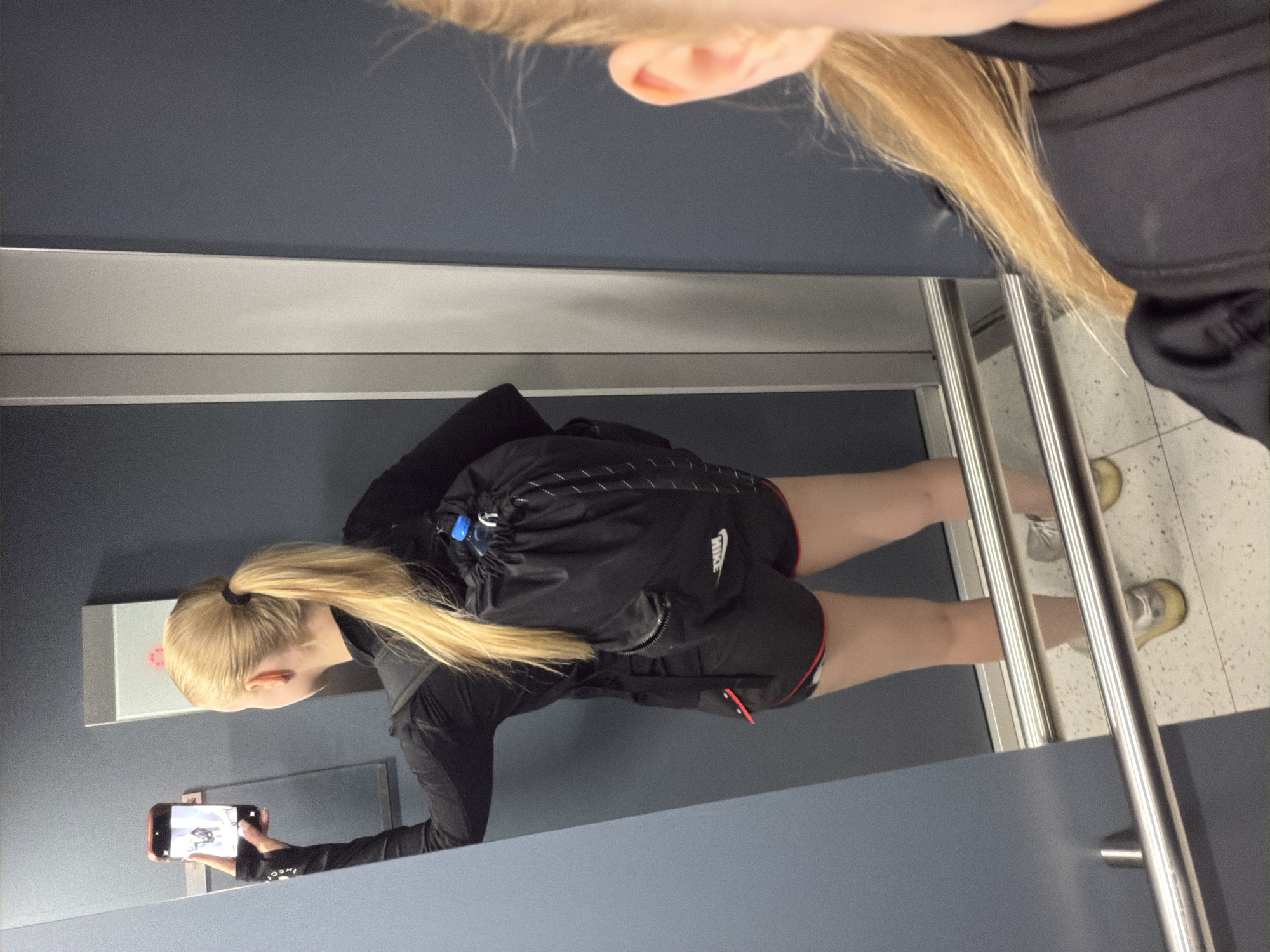 The image shows a woman with long blonde hair in a ponytail, wearing a black athletic jacket, black shorts with a Nike logo, and gold athletic shoes. She is holding a smartphone and appears to be taking a photo in a mirror. The reflection in the mirror shows a grey-toned wall with a silver accent strip, and what appears to be a light switch. Adjacent to the reflection is a stainless steel railing and a tiled floor. The location is unknown.