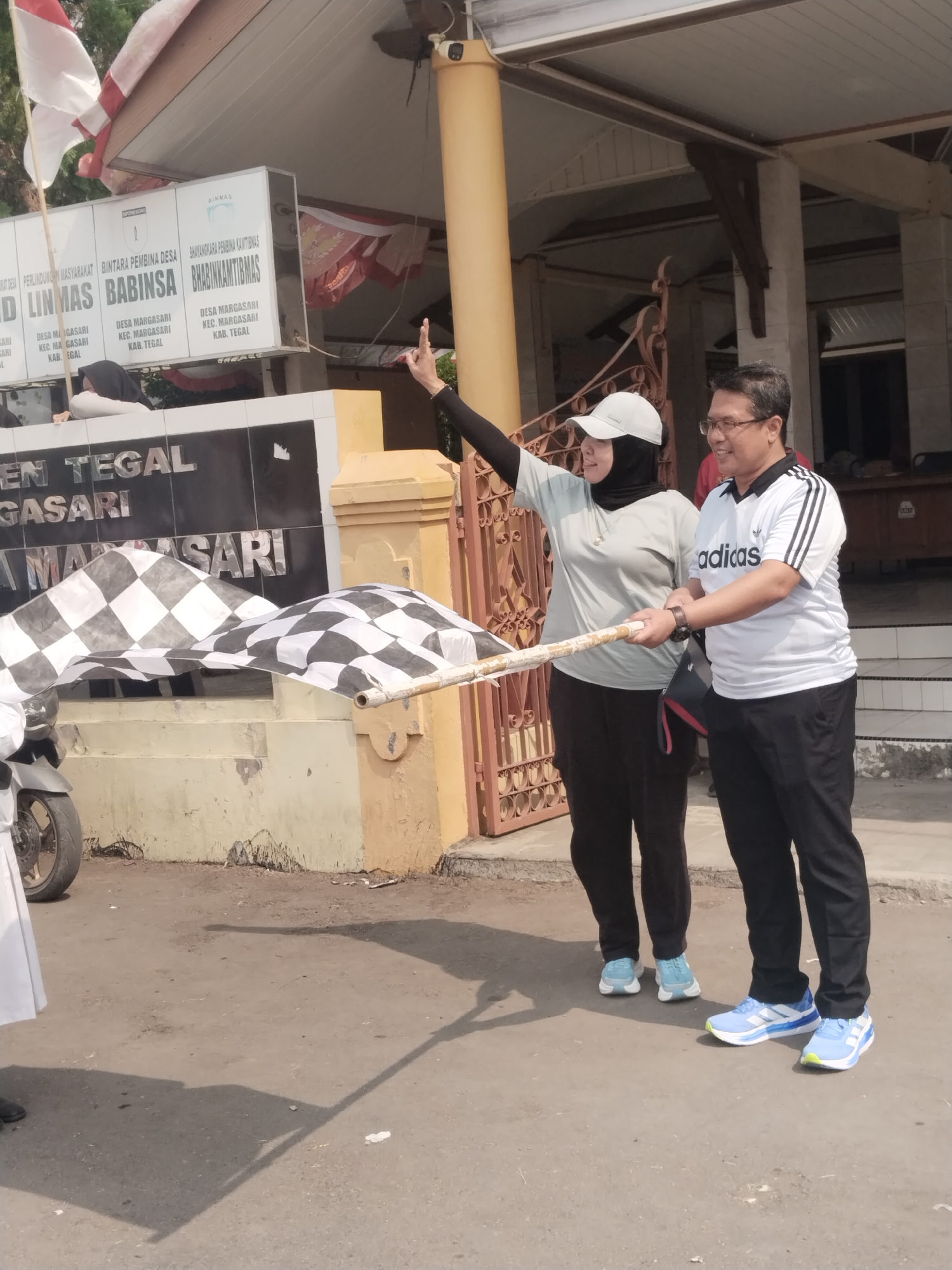 In front of a building with signage that reads "DIN TEGAL MARGAASARI" and "BINTARA PEMBINA DESA BABINSA BHABINKAMTIBMAS DESA MARGASARI KEC. MARGASARI KAB. TEGAL," two individuals, a woman wearing a black hijab and a light gray t-shirt, and a man wearing glasses and a white Adidas polo shirt, are holding a checkered flag. The flag appears to be a finish line flag. They are both looking towards the left of the frame, where the flag is being waved. The woman is pointing her left index finger upwards. In the foreground, a motorcycle is partially visible on the left side of the image. The background shows a building with a yellowish-brown facade and a white awning, and a metal gate on the right. The ground is paved with asphalt. The scene appears to be an outdoor event in Margasari, Tegal Regency, Indonesia.