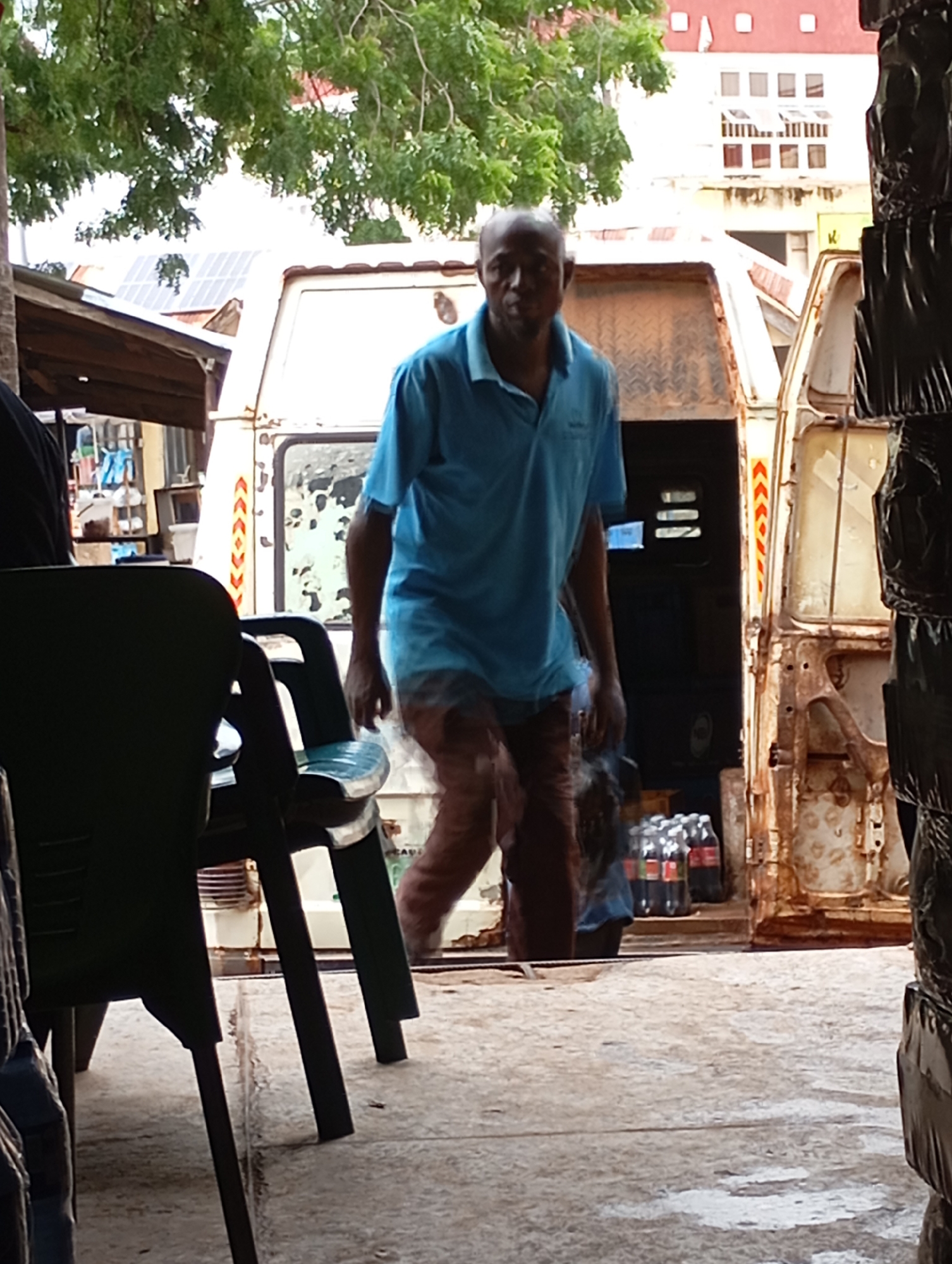 In the city of Shendam, Nigeria, a Black man wearing a blue polo shirt and brown trousers walks past a white, weathered van. He is seen from the waist down. The van is open at the back, revealing a dark interior and a few bottles of Coca-Cola on the ground. The man is partially obscured by a dark green plastic chair in the foreground. A building with red and white accents is visible in the background. The scene appears to be outdoors, with a concrete surface and a tree with green foliage overhead.