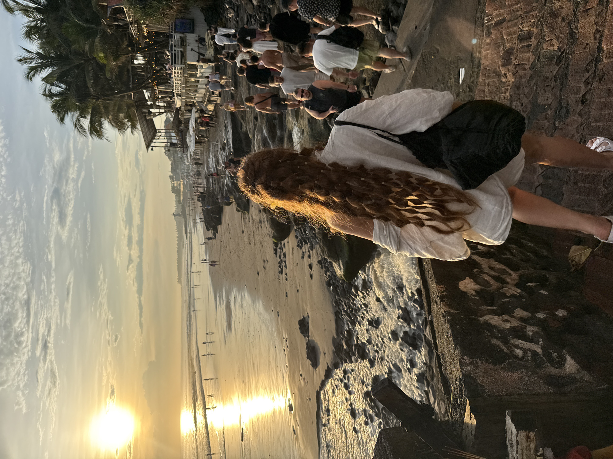 The image captures a scene at a beach in North Kuta, Indonesia. The sun is setting, casting a warm glow over the beach and ocean. A crowd of people is gathered on the beach and a raised walkway alongside the beach. A woman with long, curly brown hair stands in the foreground. She is wearing a white dress and carrying a black bag. She is positioned on a rocky edge, overlooking the beach and the crowd. Palm trees are visible in the background. The environment includes sand, rocks, and the ocean.
