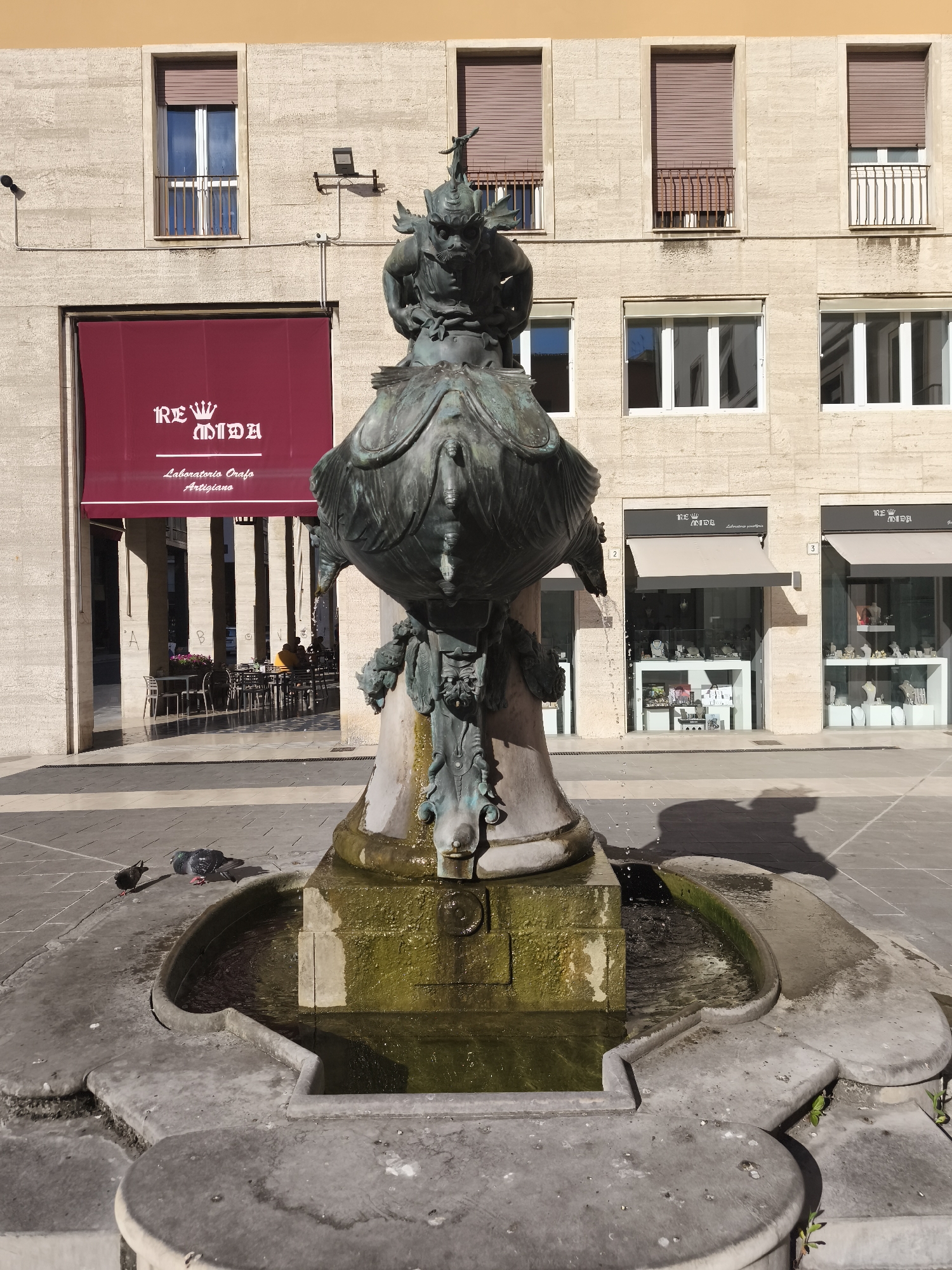 A bronze fountain, featuring a grotesque, mythical creature at its apex, stands in a paved public square. The creature has elaborate headdresses and appears to be spewing water from its mouth. The fountain's base is worn and mossy, with water flowing into a shallow pool.
In the background, a modern building features a beige stone facade. Multiple windows with brown shutters are visible on the upper floors, while the ground floor has storefronts with awnings. One awning displays a red banner with white text. Two storefronts to the right have signs that read "RE MID A" above "Laboratorio Orafo Artigiano". The awnings of these two stores are beige.
To the left of the fountain, a red awning with white text reading "RE MIDA" and "Laboratorio Orafo Artigiano" covers the entrance to a building. Through the arched opening, outdoor seating for a cafe is visible, with several patrons seated at tables.
On the paved ground in front of the fountain, two pigeons are present, one standing and another seemingly resting or pecking at the ground. The scene appears to be set in the daytime, with sunlight casting shadows on the pavement. The architecture and style of the storefronts suggest an urban European setting. The overall mood is serene and undisturbed, with the sounds of flowing water and distant urban life.