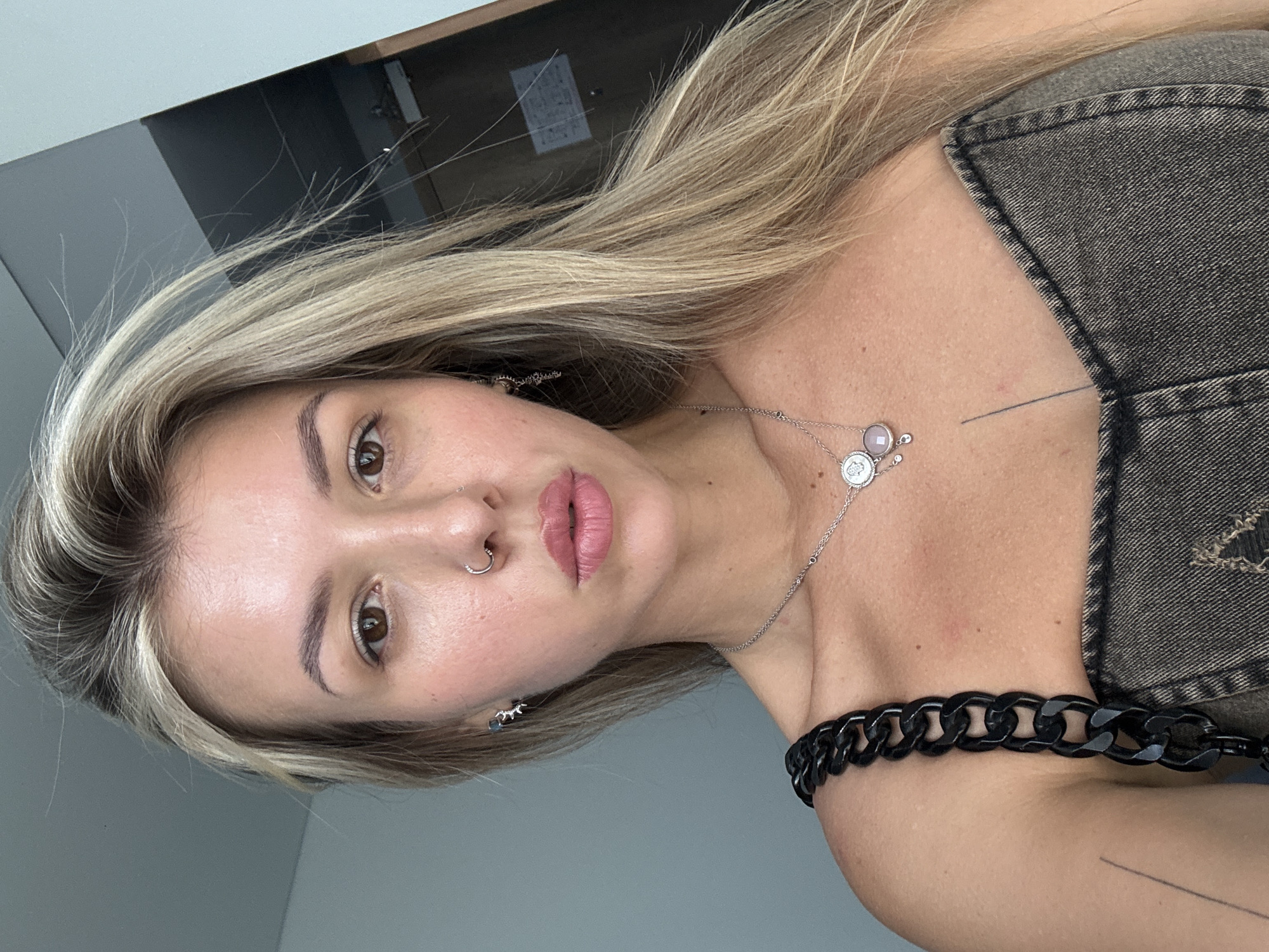 A close-up, waist-up shot shows a young, light-skinned woman with long, blonde hair and brown eyes. She is wearing a dark, strapless denim top with a thick black chain strap over her left shoulder. She has a nose ring and several necklaces, including one with a coin-shaped pendant and another with smaller charms. Her makeup is minimal, with a focus on her lips, which are lightly colored. There is a thin, vertical tattoo on her chest. The background is blurred, but appears to be an indoor setting with a light-colored wall on the left and a dark door on the right. The lighting is soft and natural.