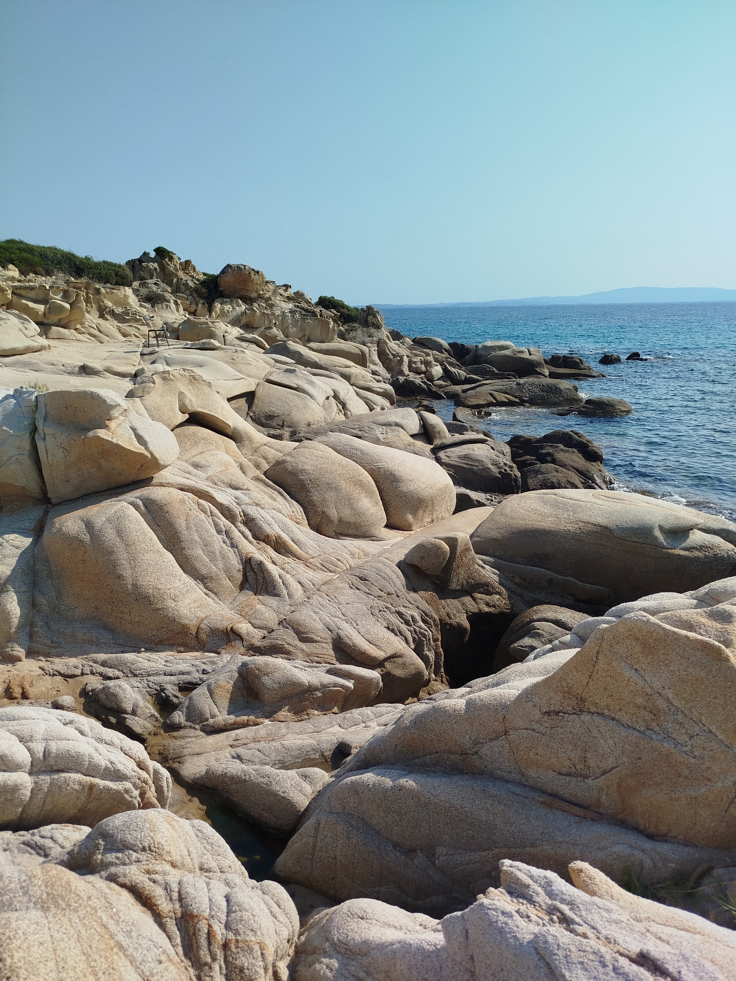 This image shows a rocky coastline with the sea in the background. The rocks are large and weathered, with rounded shapes and a light tan color. Some of the rocks have deep crevices and shadows, suggesting they are hollow or have eroded deeply. In the distance, the sea is a clear blue, with a faint horizon line where it meets the sky. The sky is a pale blue, indicating a clear day. There is no vegetation visible on the rocks themselves, but there appears to be some scrubby bushes on the higher ground in the background. A single, dark metal chair is visible on the rocky terrain in the mid-ground, adding a solitary, human element to the natural landscape. The scene is located on the coast of Sithonia Municipal Unit, Greece.