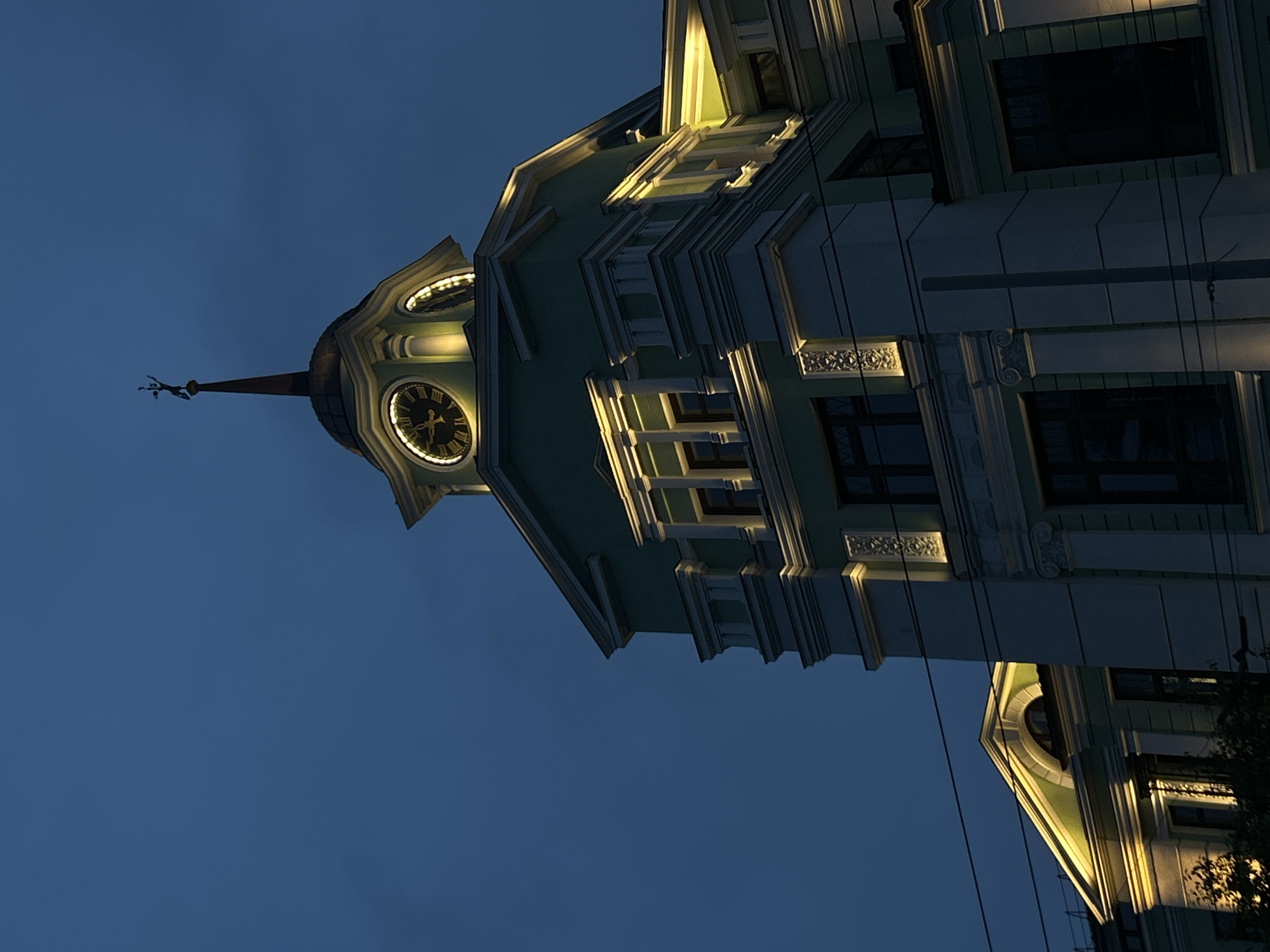 This image captures the upper portion of the Sevastyanov House (Dom Sevastyanova), a landmark building in Yekaterinburg, Russia, at dusk or night. The sky is a deep, uniform blue, indicating low ambient light conditions. The building's facade is primarily mint green with extensive light grey or white architectural embellishments, including cornices, pilasters, and decorative window surrounds.
A prominent clock tower rises from the building, topped by a rounded dome and a slender, dark spire. A metallic weathervane, shaped as a dynamic, human-like figure, is affixed to the apex of the spire. The clock tower features at least one visible circular clock face with Roman numerals, illuminated from within by a warm yellow light. Additional warm yellow accent lighting is strategically integrated along cornices, below windows, and on decorative elements across the building's visible sections, highlighting its intricate architectural details.
To the left and right of the main clock tower, other rooflines and building sections are visible, similarly illuminated. In the lower foreground, multiple thin, dark horizontal lines are discernible, consistent with overhead utility or tram wires. No individuals, vehicles, or specific activities are present in the frame.
