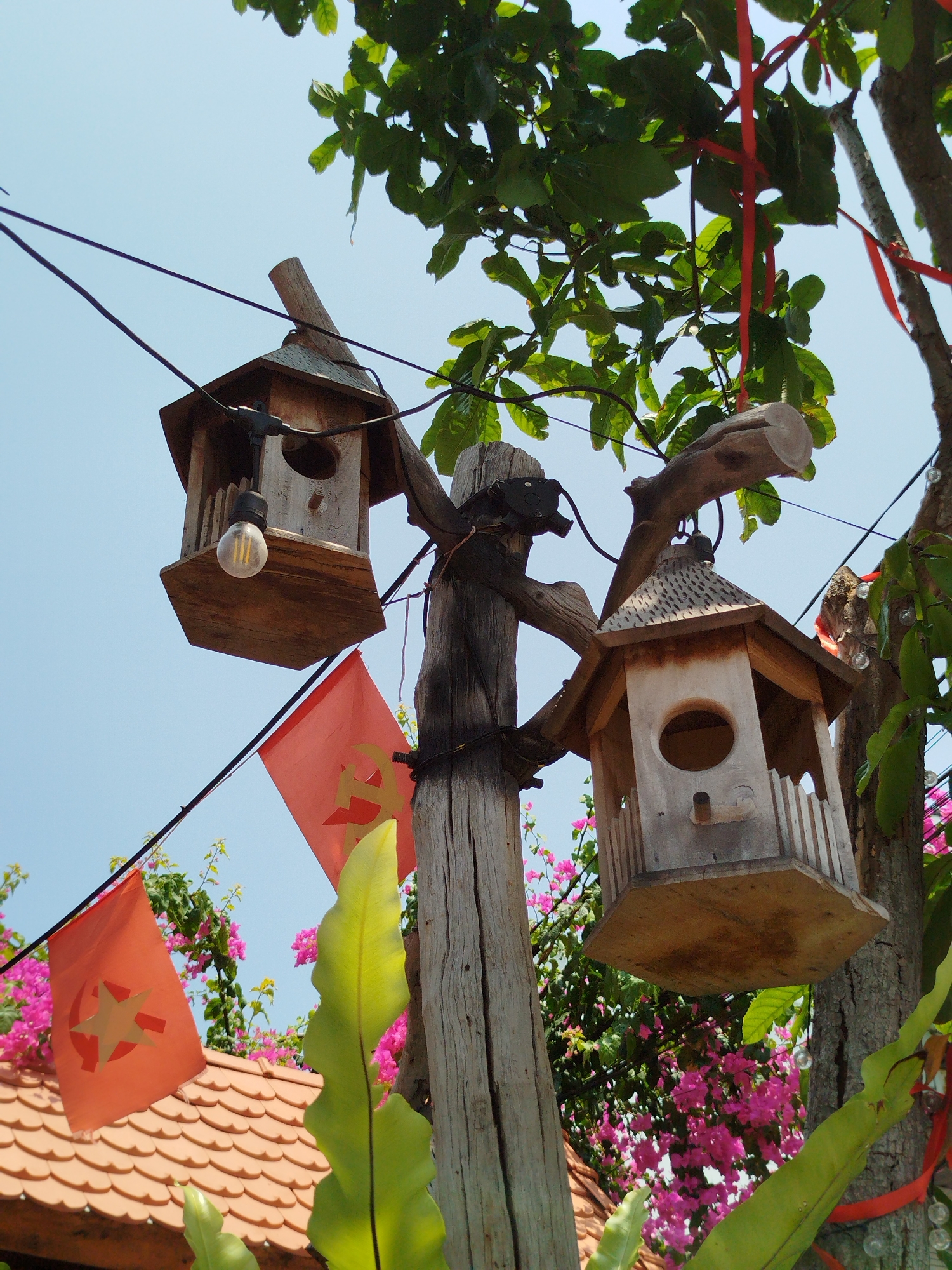 The image shows two wooden birdhouses hanging from a wooden pole and tree branches in what appears to be an outdoor setting in Ho Chi Minh City, Vietnam. The birdhouses are different designs; one is simple with a hanging lightbulb, while the other has a more decorative design with a rounded opening. Red ribbons are tied to the tree branches, and red flags with a golden hammer and sickle emblem are strung nearby. Pink bougainvillea flowers are visible in the background, along with a tiled roof. The sky is blue, indicating a sunny day.