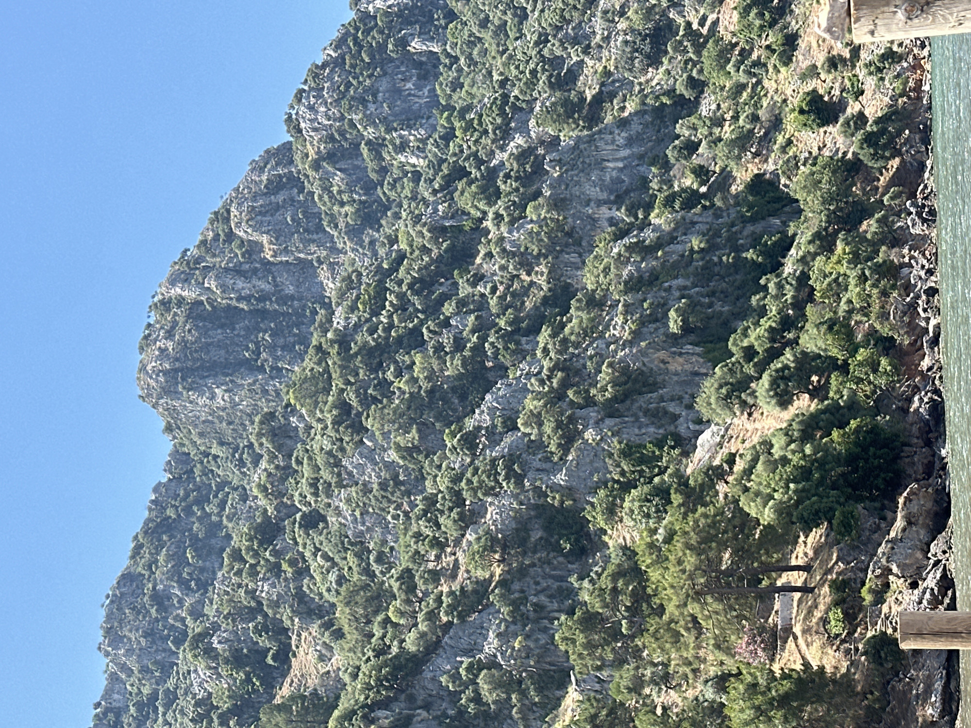 A mountain in Ortaca, Turkey is covered with rocks and trees. The mountain is gray and green. The sky is blue. The right side of the image contains a wooden beam and dark water.