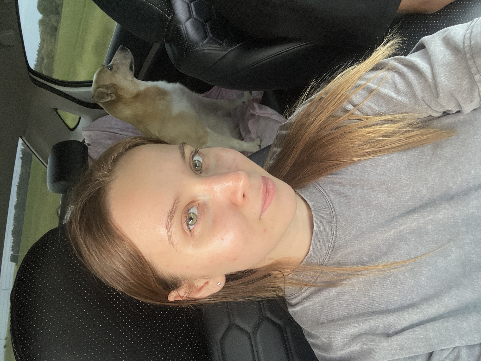 A young woman with blonde hair and green eyes is taking a selfie in the back seat of a car. She is wearing a gray sweatshirt. A light brown and white dog is sitting next to her, looking out the window. The car is on a road in a rural area, with green fields and trees visible outside. The interior of the car is dark leather. The image was taken in Povarenki, Russia.