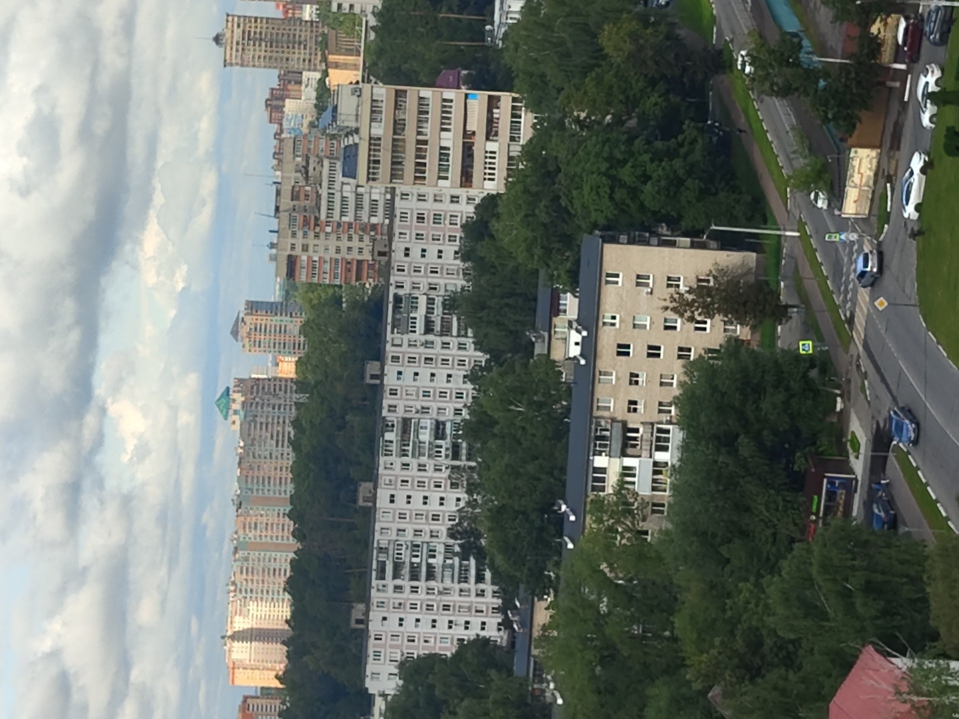 The image is an aerial view of a city in Ramenskoye, Russia, featuring a dense urban landscape with numerous apartment buildings and trees under a cloudy sky. In the foreground, a multi-story apartment building with a light-colored facade and dark window frames is visible, with trees surrounding it and a road in front. Several cars, including a blue sedan and a dark-colored SUV, are on the road. Further back, a series of taller apartment buildings with a mix of colorful facades and architectural styles dominate the cityscape. These buildings are interspersed with large areas of dense green trees, suggesting a well-treed urban environment. The sky is overcast with heavy clouds, casting a diffused light over the scene. Some smaller structures, possibly commercial buildings or kiosks, are also present along the road. The overall impression is one of a lived-in, established residential area within a city.