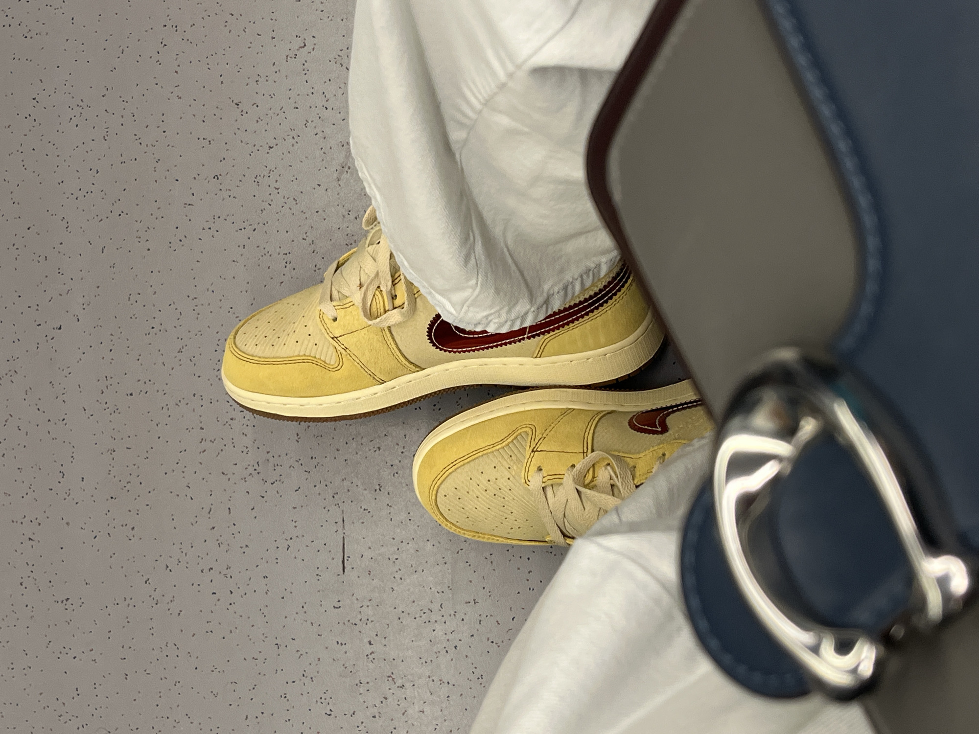 A high-angle medium shot depicts a person wearing yellow and maroon Nike Air Jordan 1 Low sneakers, white pants, and positioned on a gray speckled floor in an interior space. Adjacent to the person is a handbag or briefcase featuring a gray top, dark blue side, and a silver metal clasp. The specific location within Sofia, Bulgaria, cannot be determined from the visual information provided.