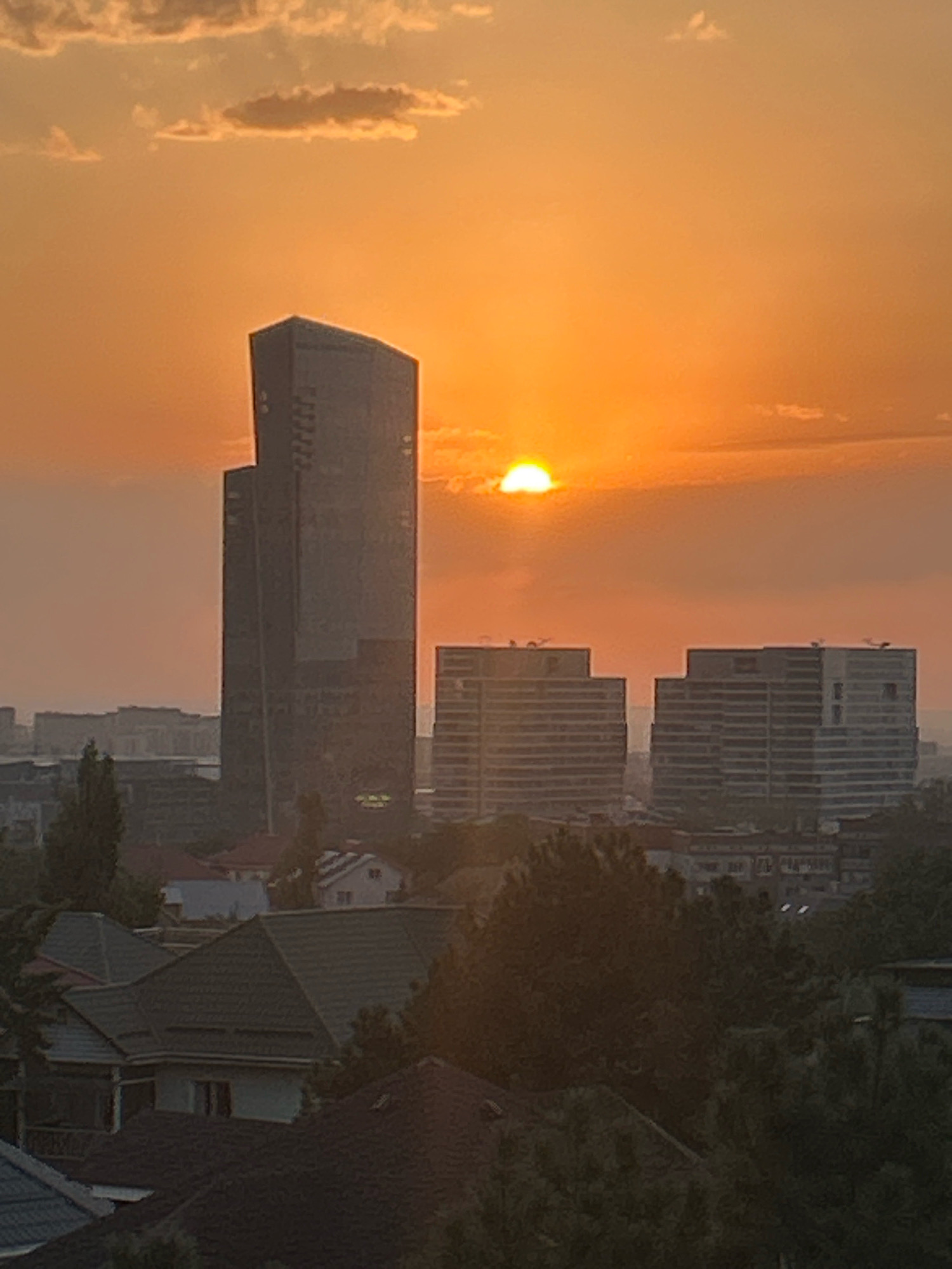 This image depicts a sunset over the city of Almaty, Kazakhstan. The sky is a vibrant orange, transitioning to yellow near the horizon where the sun is partially obscured by clouds. In the foreground, rooftops of residential buildings are visible, with trees and greenery interspersed. In the mid-ground, there are several modern buildings, including a tall, dark skyscraper with a reflective facade, and two wider, lighter-colored buildings with multiple stories. The background shows more of the city's skyline, with some buildings appearing distant and hazy. The overall impression is of an urban landscape at dusk, with the setting sun casting a warm glow over the scene.
