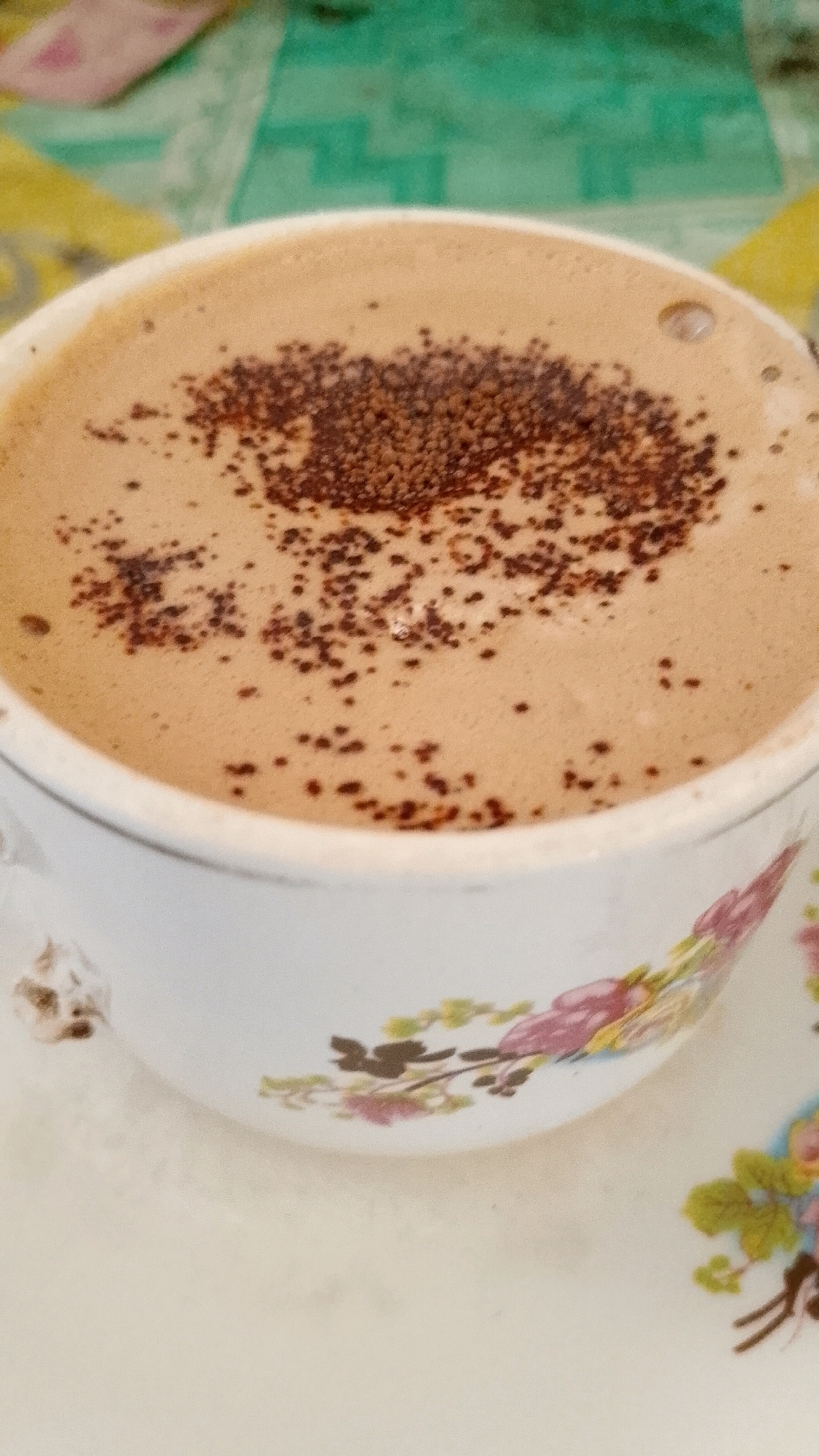 A close-up, slightly overhead shot shows a white ceramic cup filled with a foamy, light brown beverage, possibly coffee or hot chocolate. The surface of the drink is dusted with dark brown powder, scattered unevenly. A single bubble is visible on the right side of the drink's surface. The cup has a floral pattern with pink and green flowers and brown stems. The cup rests on a matching saucer. The background is blurred, showing a surface with a greenish-blue patterned textile. This scene likely takes place on a table in Mojopetung, Indonesia.