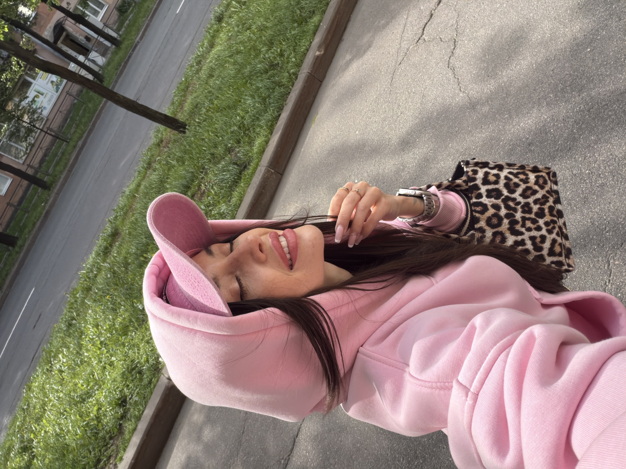A young woman is posing for a photograph outdoors in Saint Petersburg, Russia. She is wearing a pink hooded sweatshirt and a pink cap, and she is holding her hand up to her face, showing off her rings. She also wears a watch. A leopard-print bag is resting beside her on the asphalt. In the background, there is a street with grass and trees on one side and a building on the other.