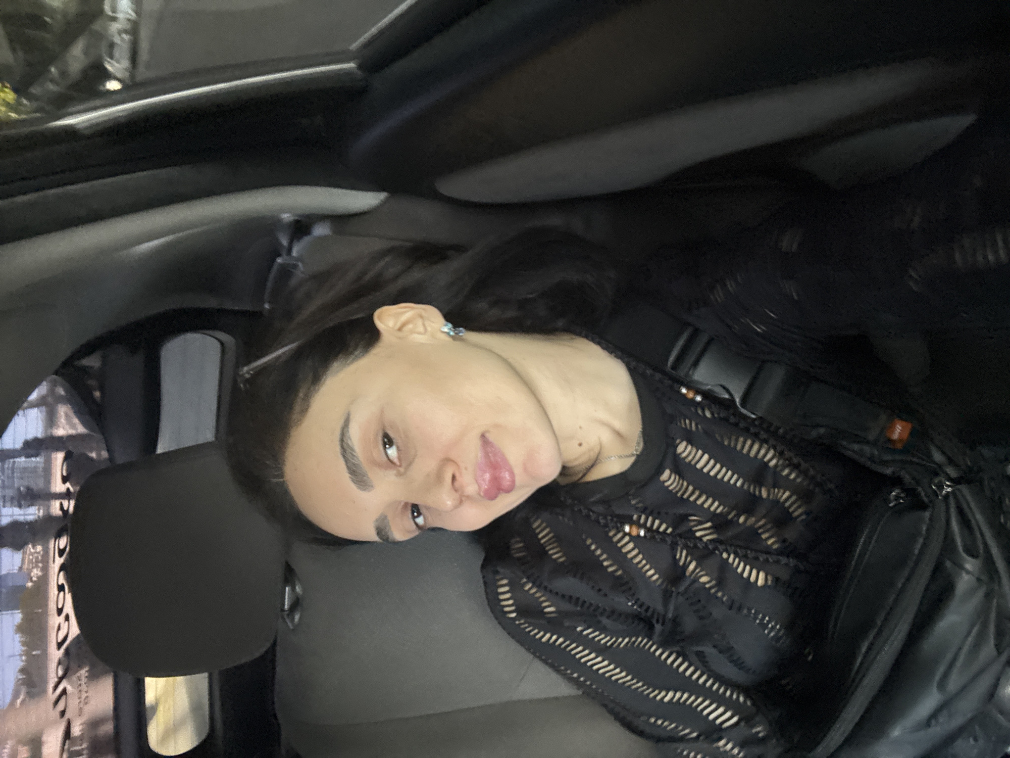 A person with long dark hair, wearing a black sheer top with horizontal stripes and a black bag, is sitting in the back seat of a car. The car is likely in the city of Tbilisi, Georgia, based on the visible cityscape outside the car window. The person is angled towards the camera and is looking at the camera.