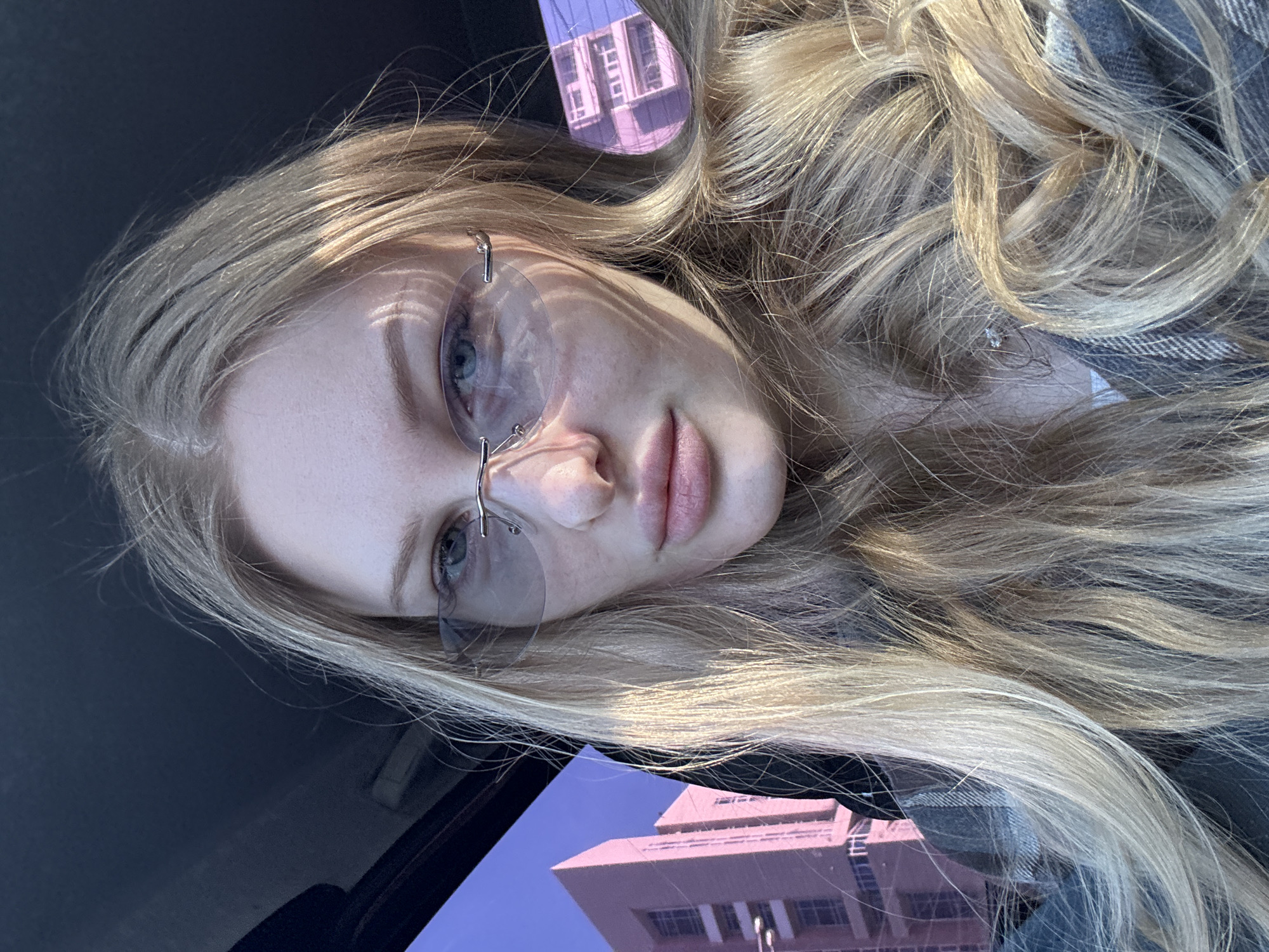 The image shows a blonde woman with blue eyes and frameless glasses. She is lying down, possibly in a car. Her head is angled towards the camera. The reflection of a pink building with multiple windows and a tall structure is visible in the car's window and possibly also in the lenses of her glasses. Based on the building's architecture, it's likely the image was taken in Perm, Russia, near the "МФЦ" building at 25 Druzhby Street. The woman is wearing a gray and white patterned shirt. The scene appears to be outdoors during the day, with sunlight illuminating her face and hair.