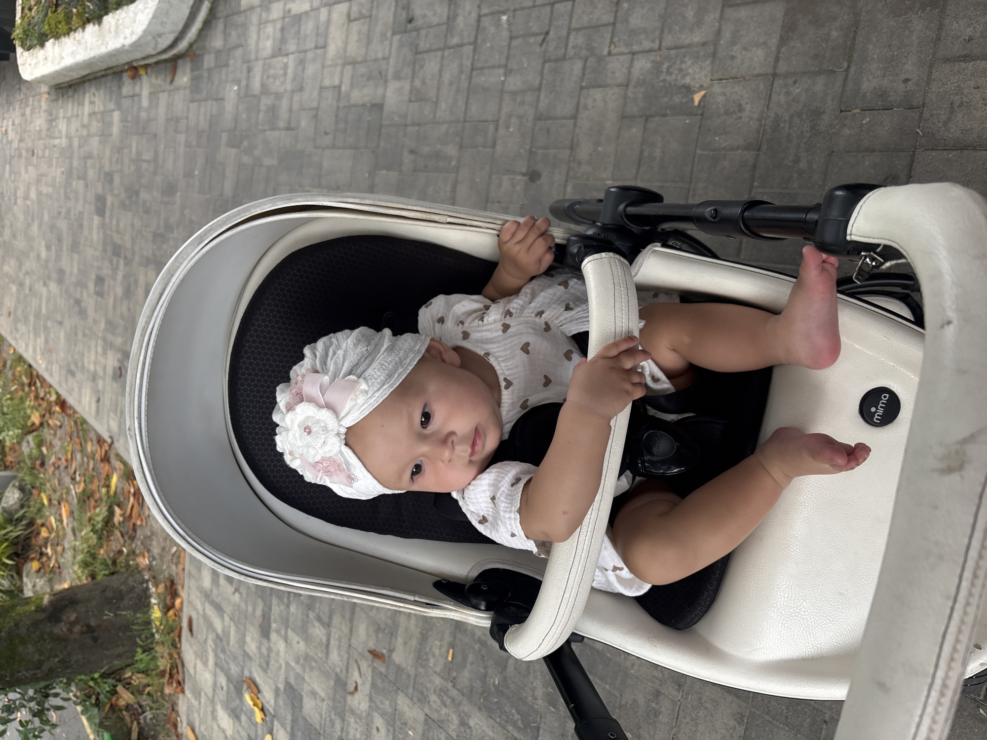 A baby sits in a white stroller on a paved walkway. The baby is wearing a white turban with a pink and white flower embellishment and a white dress with small heart patterns. The stroller has black straps and a white handle. The background shows a paved walkway with some fallen leaves and a tree. The scene appears to be in an outdoor public area, possibly a park or promenade, and the presence of palm trees, prevalent in Sochi, Russia, along with the style of paving stones and urban park design, suggests the location is the city of Sochi, Russia.