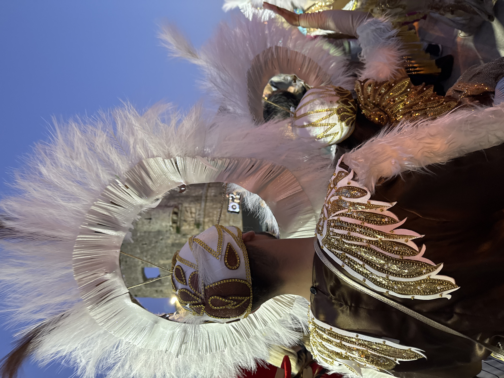 This image captures a person adorned in an elaborate, festive costume, viewed from an upward and tilted perspective. The main subject wears a striking, circular headdress made of numerous white feathers, which frames the head and neck. A decorative headpiece, primarily white and gold with intricate patterns and beadwork, covers the back of the individual's head.
The costume's garment is a deep brown, possibly satin, embellished with ornate gold and white designs that resemble wings or elaborate scrolls, adorned with sequins and beads on the shoulders and upper back. Several spherical, white and gold decorated elements are also integrated into the costume.
The setting appears to be outdoors, beneath a clear, bright blue sky, indicating daytime. In the blurred background, a stone building with arched openings is visible, suggesting an ancient or historic architectural context, consistent with locations in Budva, Montenegro. The elaborate attire strongly suggests participation in a parade, carnival, or cultural festival. There is no visible text in the image.