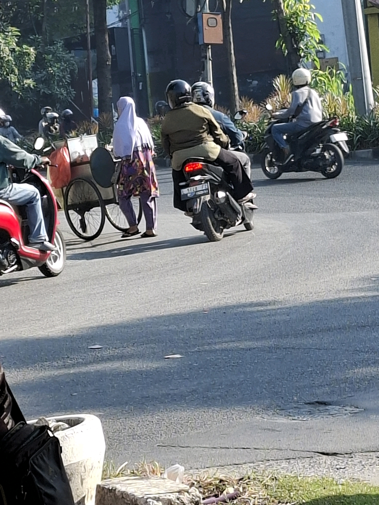 A street scene in Surabaya, Indonesia, shows several people on motorbikes and a woman with a cart on a paved road. Sunlight casts shadows on the road, indicating it is daytime.
In the foreground, a person on a red scooter is partially visible on the left. To their right, a woman wearing a white hijab and a colorful patterned skirt stands next to a cart with two wheels, one of which is large and spoked. The cart appears to be used for selling goods.
Further back, two people are riding a dark-colored motorbike. The person in front wears a green jacket and a dark helmet, while the person behind them wears a blue shirt and a dark helmet. Behind them, another person on a light-colored motorbike wears a light gray shirt and a white helmet. The motorbikes have license plates that are partially visible.
In the background, greenery and trees line the side of the road, with some buildings and a traffic signal visible further back. The overall impression is a typical street scene in a bustling Indonesian city.