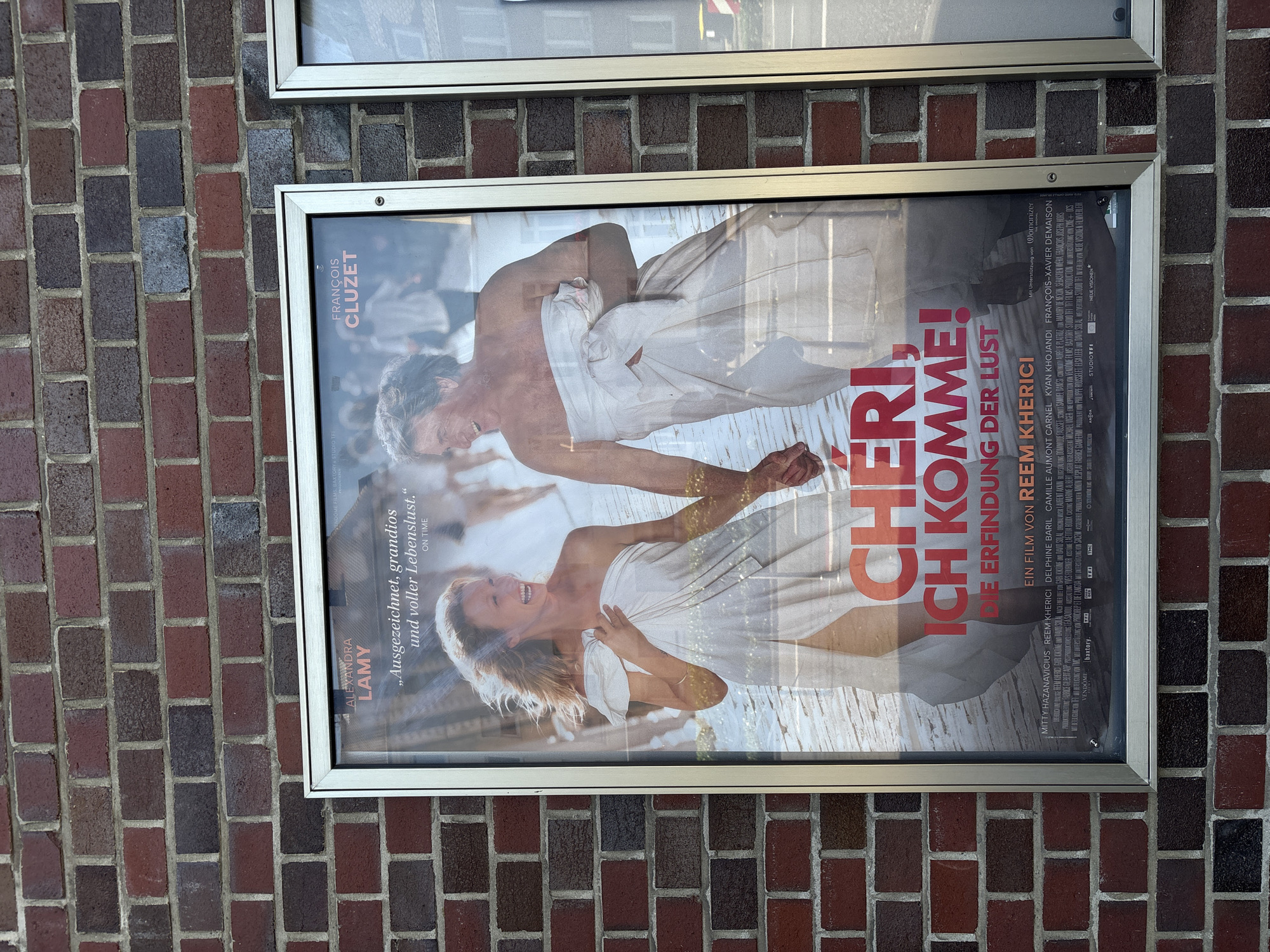 A movie poster for "Chéri, ich komme! Die Erfindung der Lust" is displayed on a brick wall in Mönchengladbach, Germany. The poster features a close-up of a man and a woman, both wrapped in white sheets, holding hands and smiling. The woman is in the foreground, laughing, while the man is slightly behind her. The background is blurred, showing other people in similar attire. The movie title and actor names, Alexandra Lamy and François Cluzet, are visible at the top of the poster. The bottom of the poster contains credits and logos. The brick wall is made of red and dark brown bricks. The poster is framed by a silver metal border.