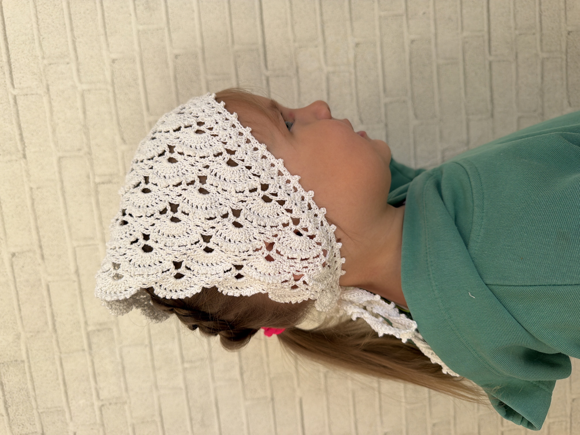 A child wearing a white crocheted bonnet and a green shirt is looking upwards. The background is a light-colored brick wall. The child has brown hair pulled back into a ponytail with a pink hair tie. The location is indeterminate and there is no specific indication it is in the city of Chaikovsky, Russia.