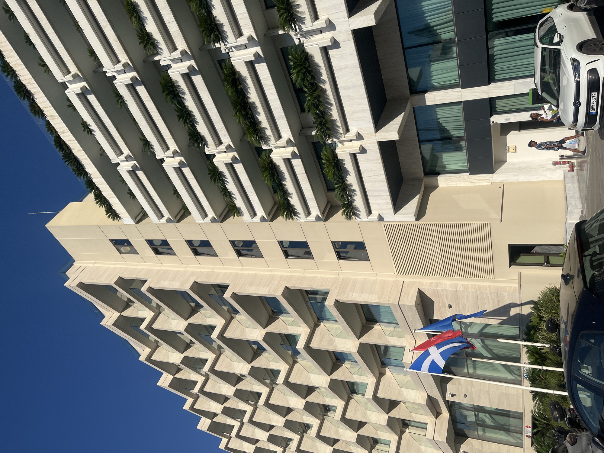 A low-angle, full shot captures two modern, beige buildings in Kallithea, Greece, against a clear blue sky. The building on the left features a facade with a repeating pattern of angled balconies, each with glass railings and some containing greenery. The building on the right is characterized by multiple horizontal balconies adorned with lush green plants. Three flags, blue and white, red, and blue, are prominently displayed on flagpoles in front of the buildings. In the foreground, a white SUV with a visible license plate (BMK-6672) is parked, and a dark car is partially visible on the left. Two individuals, a man and a woman, are walking towards the entrance of the building on the right, with the woman appearing to be holding bags. The environment appears to be a street or a plaza in front of a commercial or residential complex.
