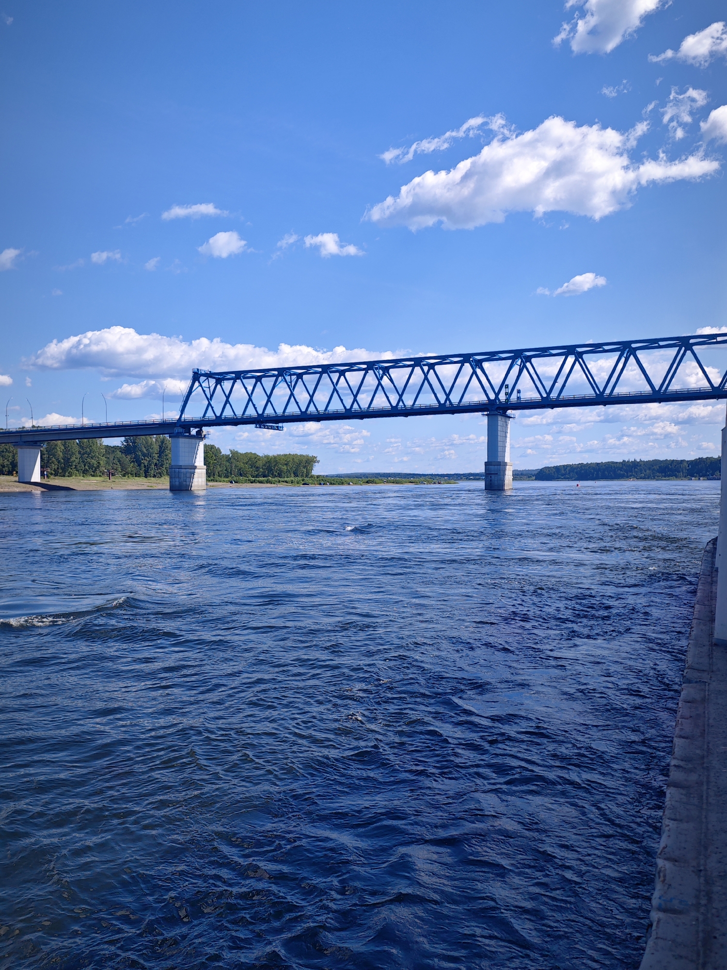 The image shows a wide, blue river with choppy water under a bright blue sky with scattered white clouds. A large blue truss bridge spans the river from left to right. The bridge is supported by concrete pillars. On the far bank, a line of green trees is visible. On the right side of the image, a concrete embankment or walkway runs along the edge of the water. The scene appears to be a sunny day, likely in Abalakovo, Russia, given the presence of the bridge which is a prominent landmark there. There are no people visible in the image.