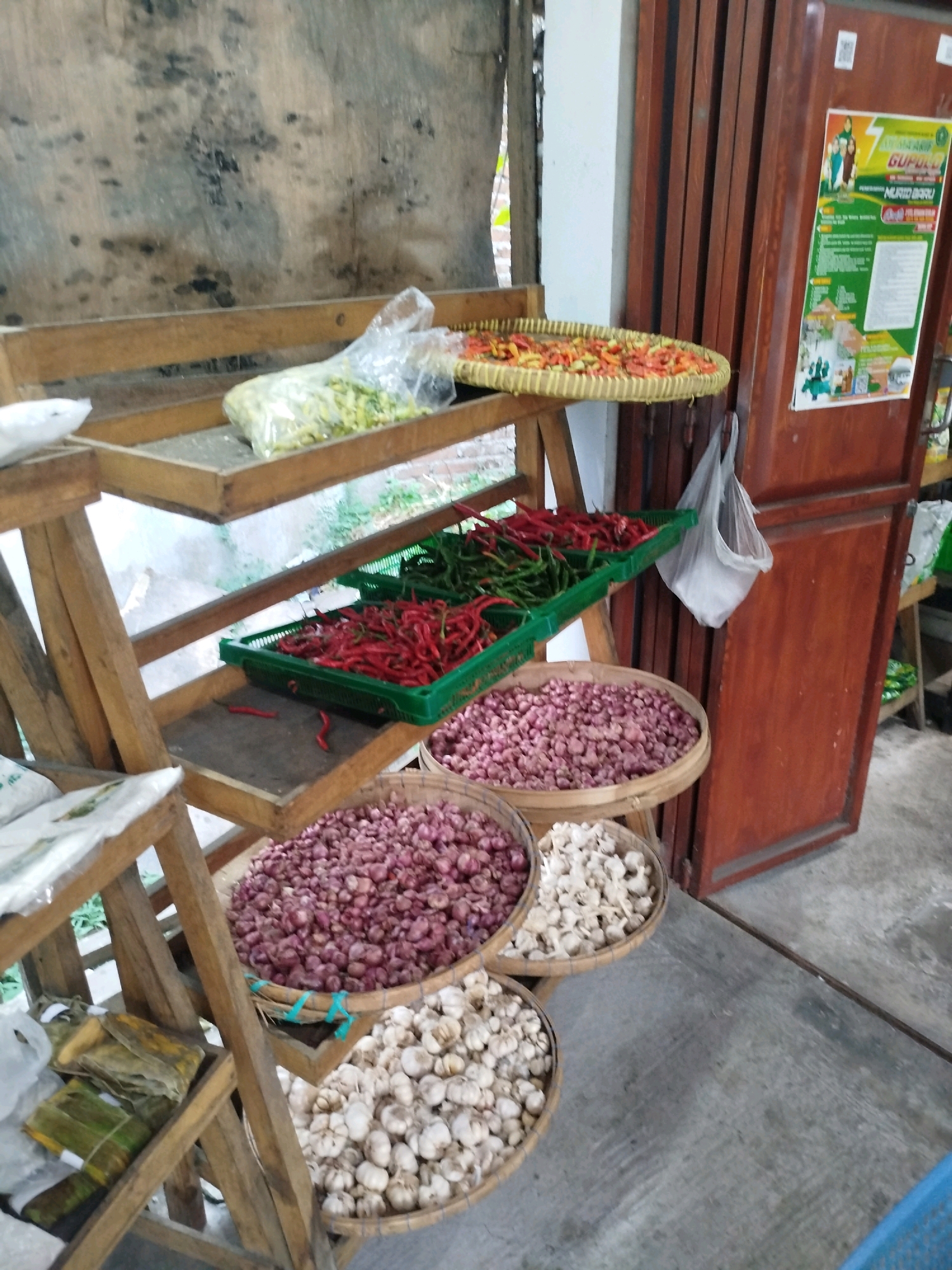This is a close-up, eye-level, medium shot of a rustic market stall in Cekok, Indonesia. The stall is made of weathered wood, with multiple shelves displaying various fresh produce.
On the top shelf, a clear plastic bag contains what appears to be fresh ginger. To its right, a woven bamboo tray is filled with red chilies. The second shelf holds two green plastic baskets, one filled with red chilies and the other with green chilies. Below these, a large woven basket is overflowing with red onions, and next to it, another basket contains garlic. Further down, wrapped items in banana leaves, secured with string, are visible.
In the background, a tall, dark wooden door stands to the right, adorned with a poster. A white plastic bag hangs from a hook near the door. The floor is a concrete surface. The overall impression is one of a local market selling fresh produce, likely for culinary use. There are no people visible in the image.