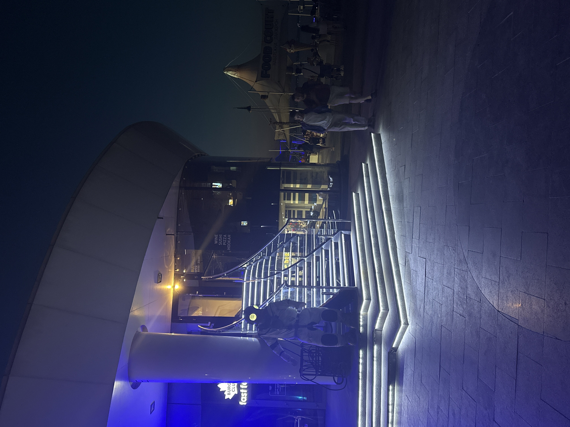 The image depicts a nighttime scene at the Odesa Sea Port's entertainment district, specifically near the "Porto Franko" complex. In the foreground, a person in a full-body astronaut costume stands on a paved area illuminated by blue lights and a well-lit staircase. The staircase leads up to a building with glass windows and signs indicating it is a lounge and restaurant. One sign reads "Fast Food." Further in the background, a large white tent structure with "FOOD COURT" written on it is visible, along with other smaller tents and structures, suggesting a bustling area with various food vendors and entertainment options. Two men are walking away from the viewer in the middle ground, and other people can be seen in the distance. The sky is dark, indicating it is nighttime. The overall atmosphere is vibrant and modern, with a focus on entertainment and dining.