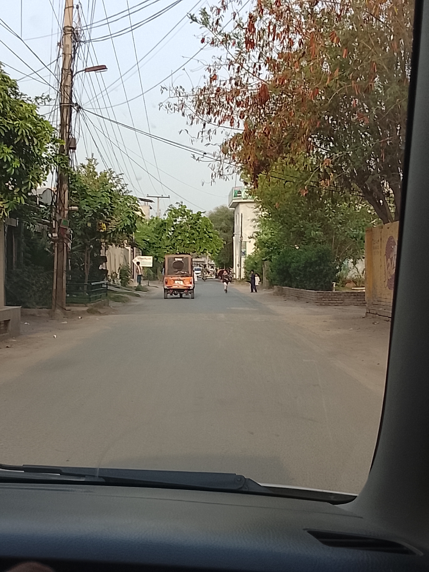 The view is from inside a car looking out onto a street in Nasheman Colony, Pakistan. The road is paved and stretches straight ahead with a slight curve in the distance. On the left side of the road are buildings and green trees, some with a sign. A utility pole is visible, with many electrical wires strung across the street. On the right side, there are more buildings and trees, including one with reddish-brown seed pods hanging down. A small orange vehicle is driving on the road. Several people are on the side of the road to the right. The sky is overcast and muted.