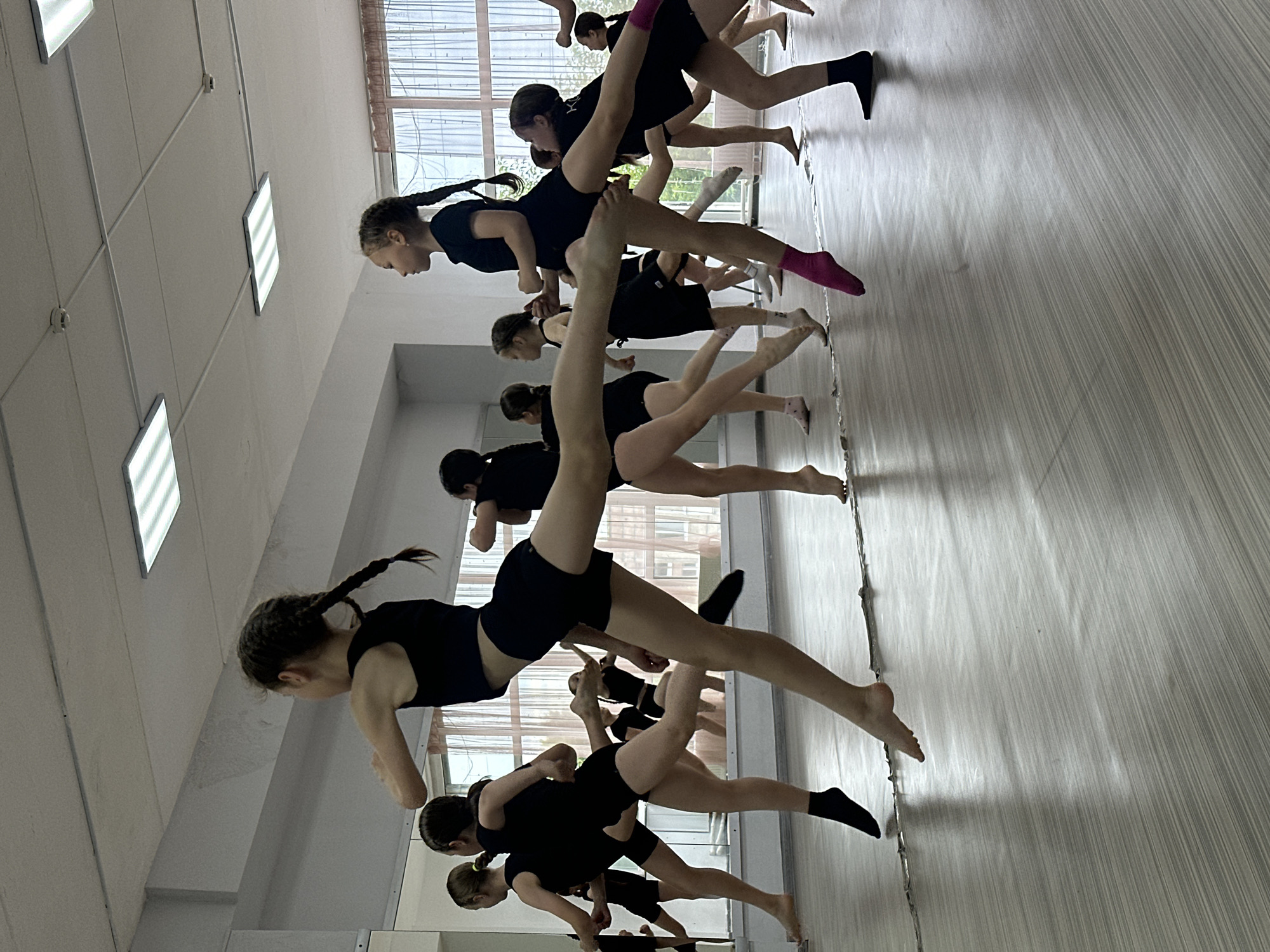 This media file captures a group of young women in a dance studio, likely in Votkinsk, Russia. The women, dressed in black leotards and shorts, are engaged in a dance exercise, possibly a rehearsal or class. Their poses suggest dynamic movement, with legs extended and bodies angled. The setting appears to be a well-lit room with a mirrored wall, reflecting the dancers and the space. Fluorescent lighting panels are visible on the ceiling, and a large window with blinds offers a glimpse of an outdoor, possibly daytime scene. The overall mood seems focused and disciplined, typical of a dance practice. The image is oriented vertically, with the dancers appearing to move from bottom to top in the frame.
