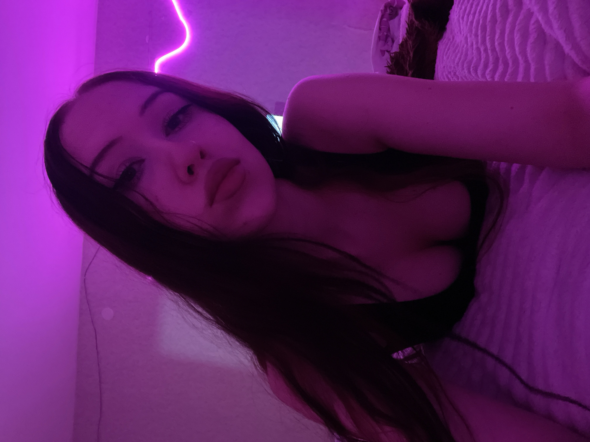 A young woman with long, dark hair is lying on a bed in a room illuminated by purple LED lights. She is wearing a dark top that reveals her cleavage. Her face is turned towards the camera, with her lips slightly parted. The room appears to be a bedroom, with a textured bedspread and a neon light strip on the wall. There is no indication of the location being Votkinsk, Russia.