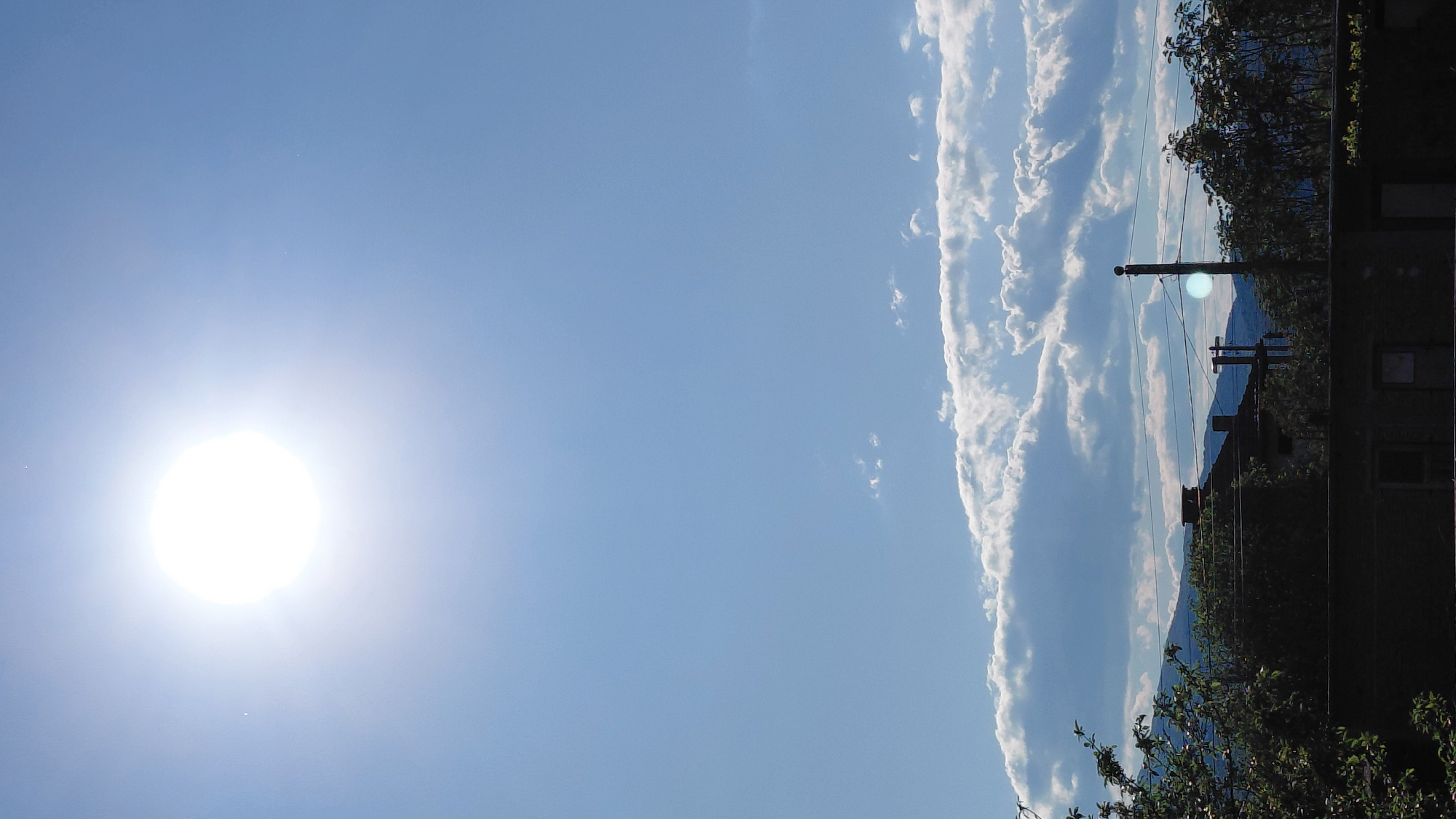 This image captures a bright, sunny outdoor scene, presented in a portrait orientation but effectively a landscape rotated 90 degrees counter-clockwise. The majority of the frame is dominated by a clear, vibrant blue sky. On the left side of the sky, a brilliant, circular sun is prominently featured, emitting an intense white light that creates lens flare and overexposure in that area.
A distinct band of white, fluffy cumulus clouds stretches across the sky on the right side of the frame (which would be horizontally across the middle of the sky if the image were rotated correctly). These clouds are well-defined and bright against the blue backdrop.
In the lower portion of the image (the right side when correctly oriented), a distant landscape of rolling mountains or hills is visible, appearing somewhat hazy and blue. In the immediate foreground on this side, dark silhouettes of dense tree foliage and branches are prominent. A utility pole, with multiple wires extending across the scene, stands out against the distant hills and sky. Parts of a dark building or structure with visible windows are also discernible on the far right edge, mostly shrouded in shadow.
The time of day appears to be late morning or early afternoon, judging by the high position and intensity of the sun. The weather is clear and sunny with these specific cloud formations. No specific activity or event is taking place; it's a static landscape view. There is no visible text in the image.