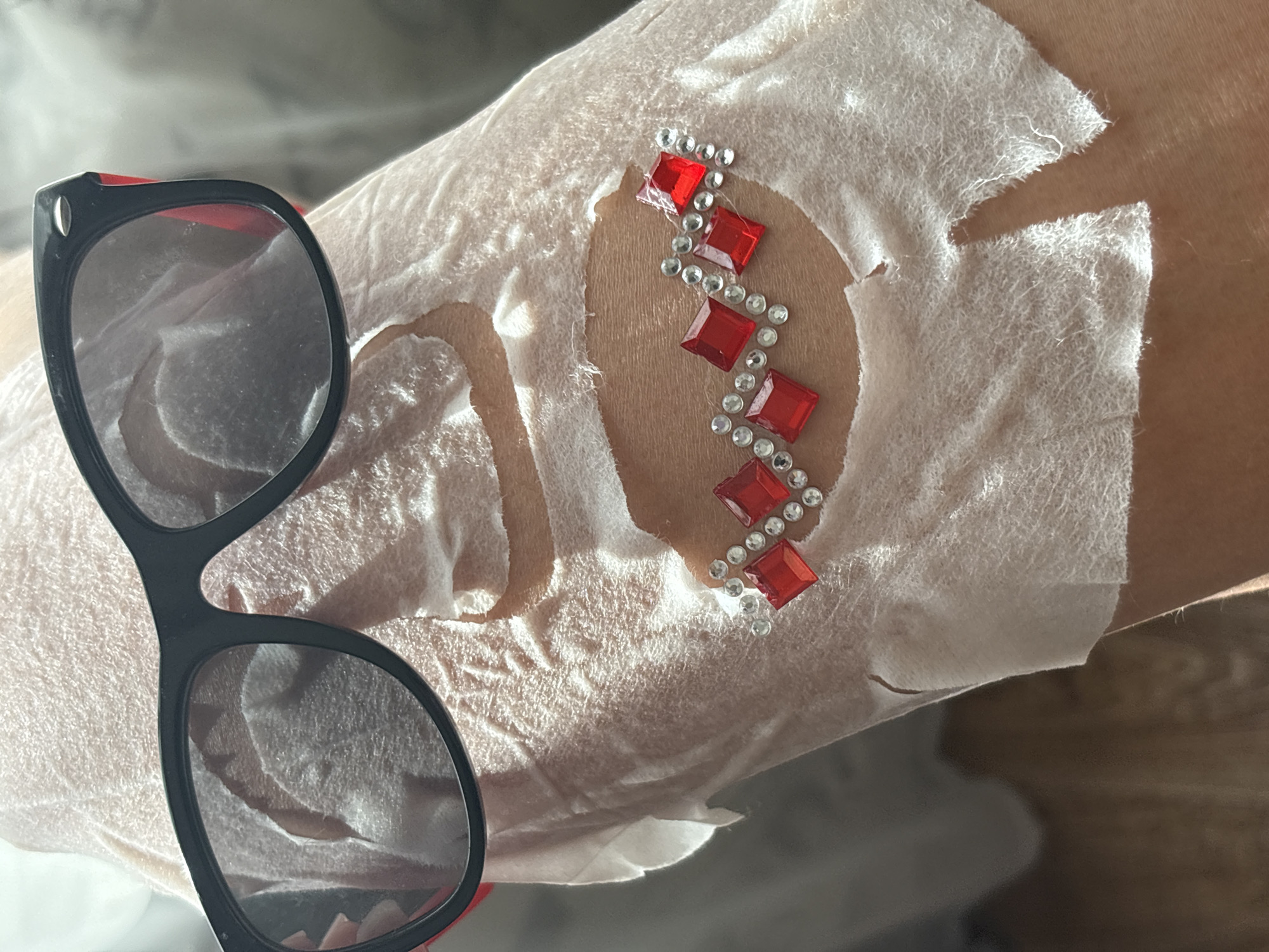 A close-up shot captures a person's arm or leg, partially covered by a piece of white, non-woven material, possibly a disposable wrap or fabric, in an indoor setting in Chaikovsky, Russia. A cutout in the material reveals skin, which is adorned with an elaborate design of red square-shaped rhinestones and smaller clear/silver round rhinestones. The rhinestones are arranged in a zig-zag pattern along the exposed skin. To the left, a pair of black-framed sunglasses with subtle red accents on the arms rests on the white material. The lenses of the sunglasses reflect a bright, sunlit indoor environment, suggesting it is daytime. The white covering material shows torn or frayed edges around the skin cutout. The activity depicted appears to be the application of temporary body art or a beauty treatment involving skin decoration. No text is visible.