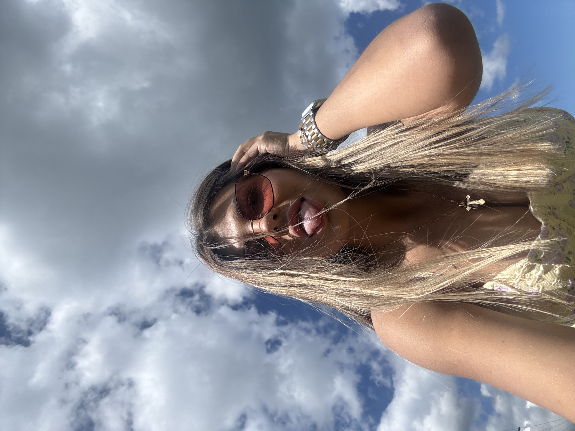 In a close-up, low-angle selfie, a young woman with long, blonde hair is pictured outdoors against a backdrop of a dynamic sky with both blue patches and grey clouds. She is wearing rose-tinted sunglasses and a yellow floral top, with a cross pendant necklace visible. Her right hand is raised, touching her hair, and her tongue is sticking out. A silver and gold watch adorns her left wrist. The image does not provide any specific details to identify a location in the city of Пастухово, Russia.