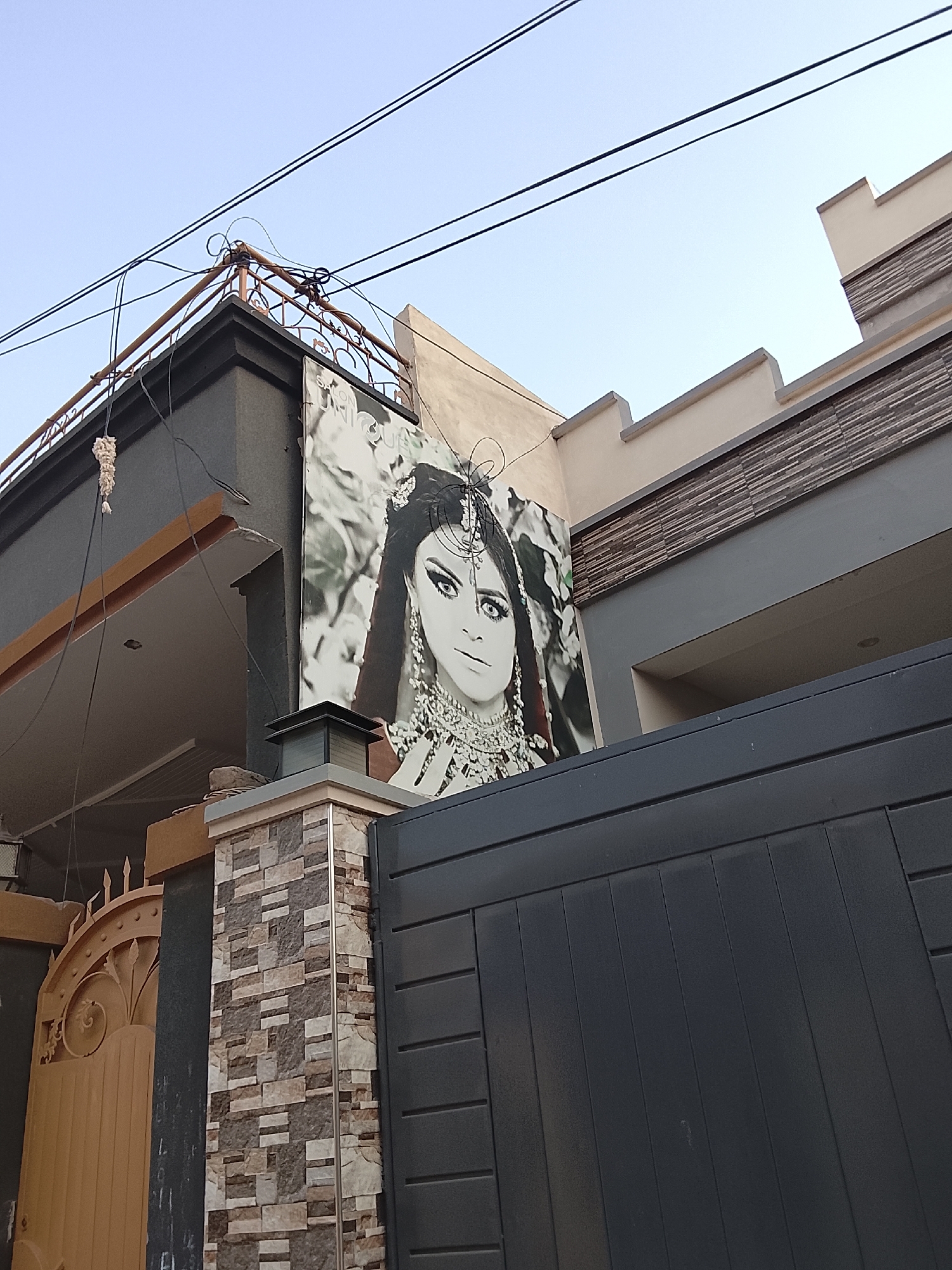 The image captures a building exterior in the city of Unknown, Unknown. The building is painted in various shades of gray and beige, with a decorative stone accent. A large black and white advertisement is affixed to the building's facade, depicting a woman adorned with jewelry. Below the advertisement, a section of a dark gray gate is visible. Adjacent to the gate is a pillar decorated with a patterned stone design. To the left, a glimpse of a golden-colored gate can be seen. Above the building, several electrical wires intersect against a light blue sky.
