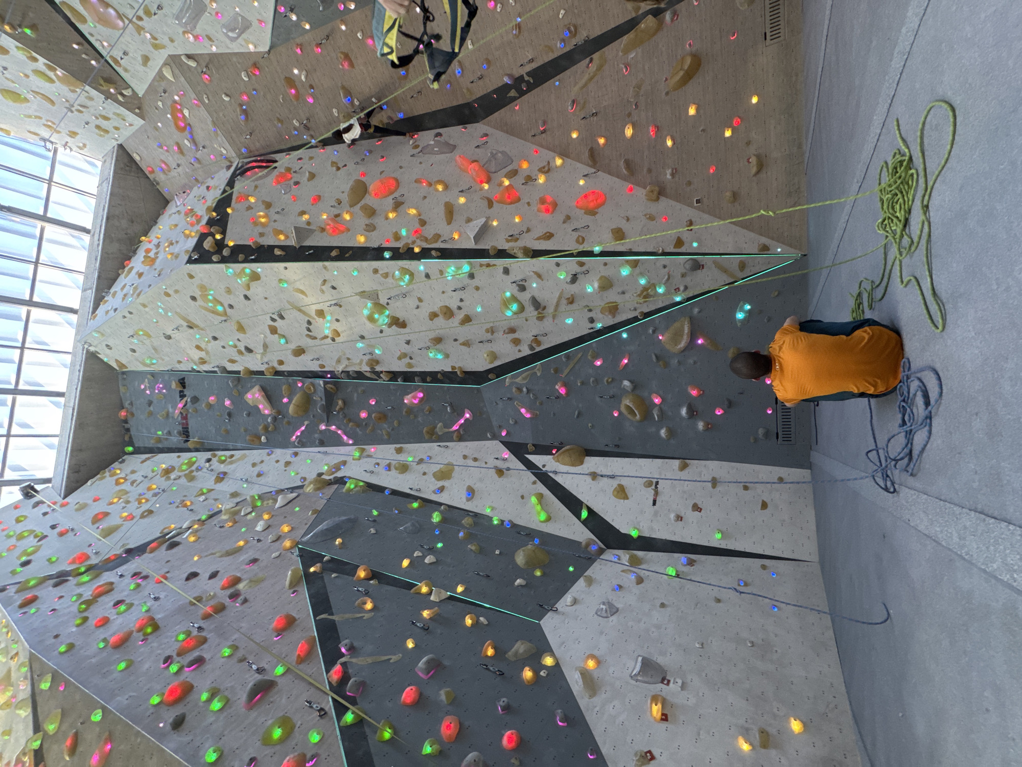 This image captures an dynamic scene inside a modern indoor climbing gym, featuring a vast and complex artificial climbing wall. The perspective is looking upwards, showcasing the height and multifaceted structure of the wall.
Two individuals are present in the frame. On the ground, situated on the right side of the image, a person wearing a bright orange shirt and dark pants is seated, seemingly belaying. A large coil of green climbing rope rests beside them, with the rope extending upwards towards the wall. Higher up on the wall, partially visible, another climber is engaged, wearing a yellow and black harness, indicating they are actively ascending or preparing to.
The climbing wall itself is a prominent subject, constructed from numerous geometric panels in shades of light grey, dark grey, and white. It is densely covered with a wide array of climbing holds, which are notable for their vibrant, translucent colors including red, orange, yellow, green, blue, and pink/purple. Many of these holds appear to be illuminated, casting colorful glows onto the wall surface, creating a visually striking effect. The wall features diverse angles, including significant overhangs, suggesting challenging climbing routes.
Natural light floods into the gym from a large window or skylight structure visible on the far left, indicating it is daytime. The overall atmosphere is bright and energetic. There is no clearly legible text visible in the image.