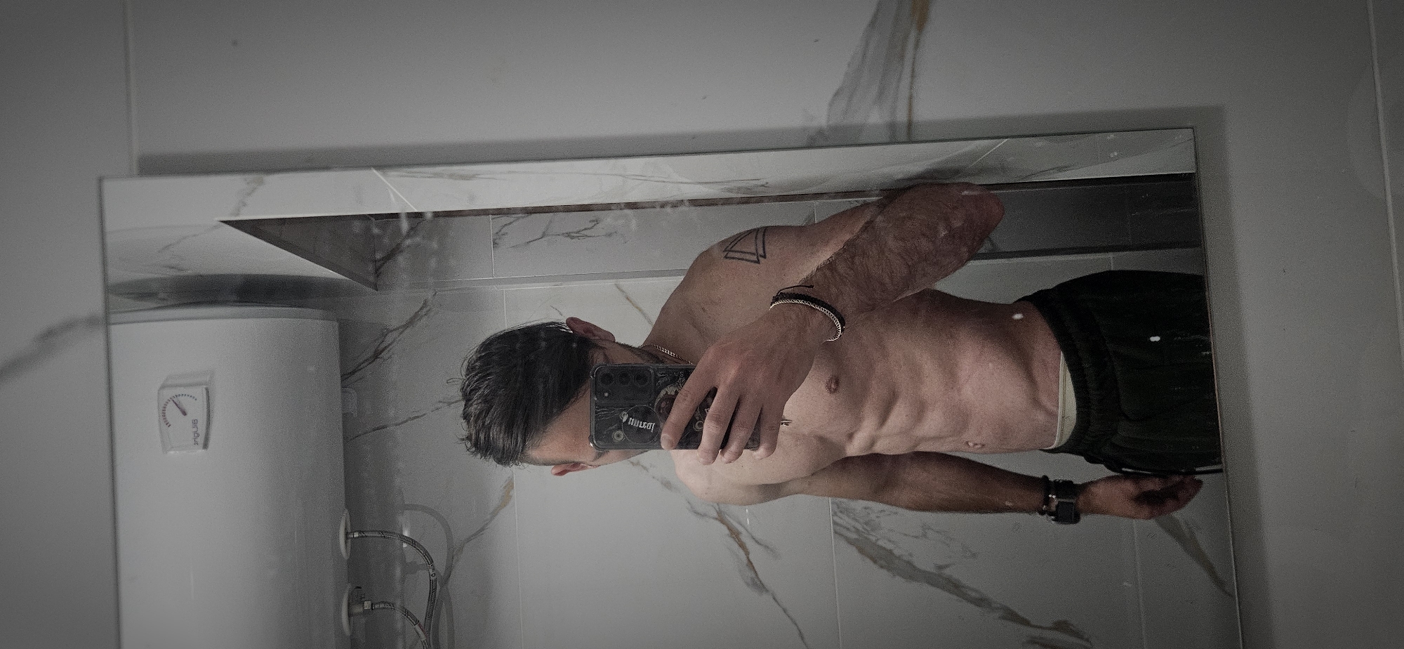 A man is reflected in a bathroom mirror. The bathroom features white walls with a marble-like pattern and a white water heater. The man is taking a photograph of himself, as his hand and phone are visible in the reflection. He has short hair and is wearing dark shorts. The man is shirtless and muscular, with a tattoo on his arm. The image appears to have been taken indoors. There is no specific information indicating the location of the bathroom in Plovdiv, Bulgaria.