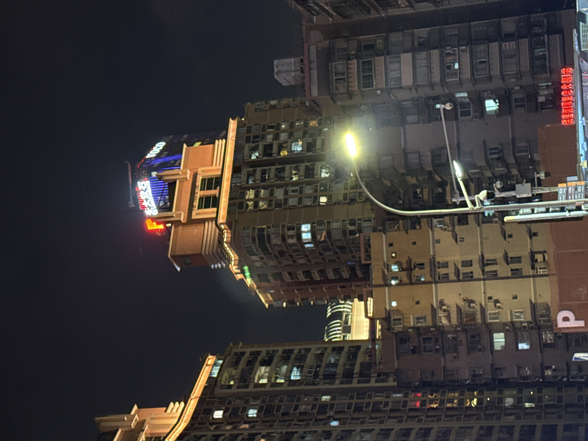 The image shows a nighttime view of a city street in Yuzhong District, China, featuring tall buildings illuminated by artificial lights. The buildings are densely packed, with visible windows, balconies, and air conditioning units. One building in the center of the frame has illuminated signage at its top, including the word "VOCO" and other unreadable text. A streetlamp with a bright bulb arches across the foreground, casting a glow on the scene. Several security cameras are visible on poles and the side of buildings. In the lower part of the image, a red sign with Chinese characters is visible, and a large white letter "P" is displayed on a brown surface. The sky is dark and appears to be cloudy or hazy.